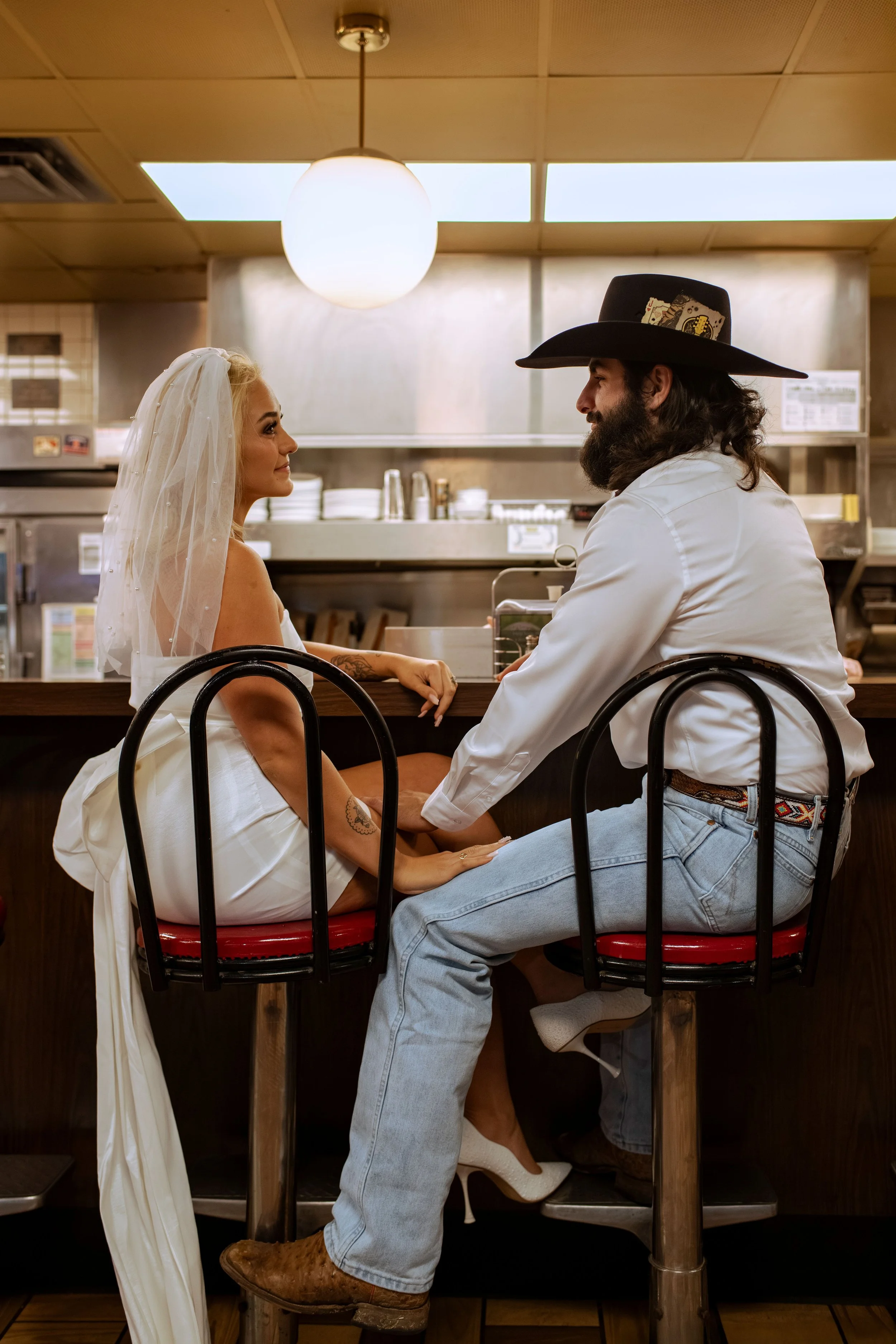A bride and a man with a beard and long hair, wearing a wide-brimmed hat, are sitting at a counter in a restaurant, holding hands and looking into each other's eyes.