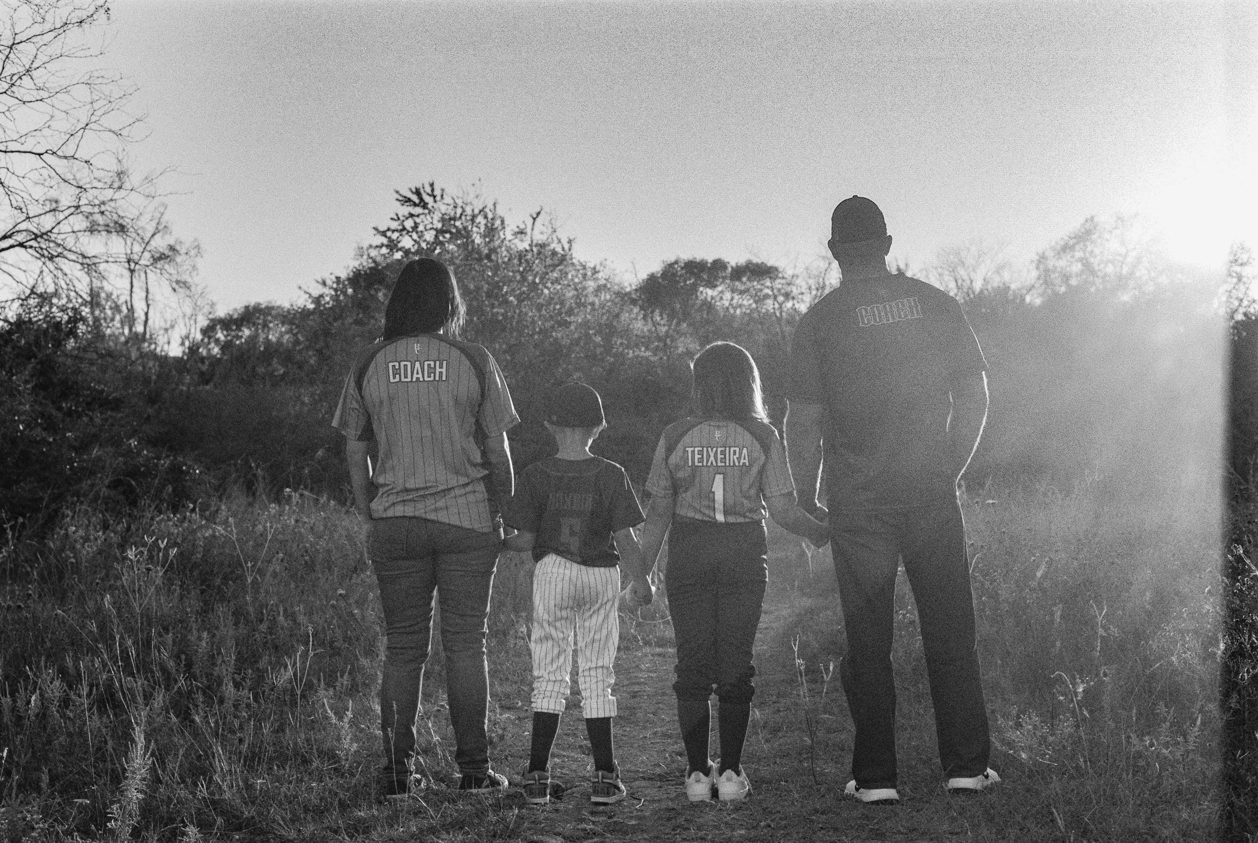 A family of four walking together outdoors during sunset, holding hands, with two children wearing baseball uniforms and two adults wearing shirts labeled 'Coach' and 'Coach'.