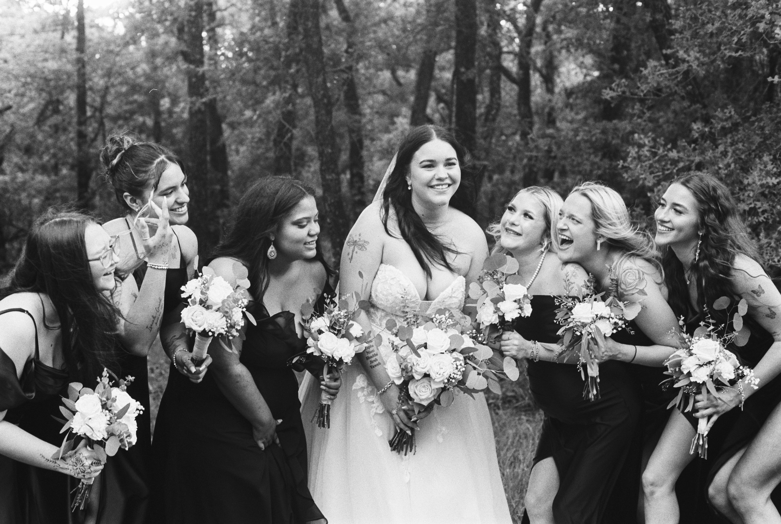 Black and white photo of a bride with dark hair smiling, surrounded by bridesmaids in black dresses holding bouquets, in a wooded outdoor setting.
