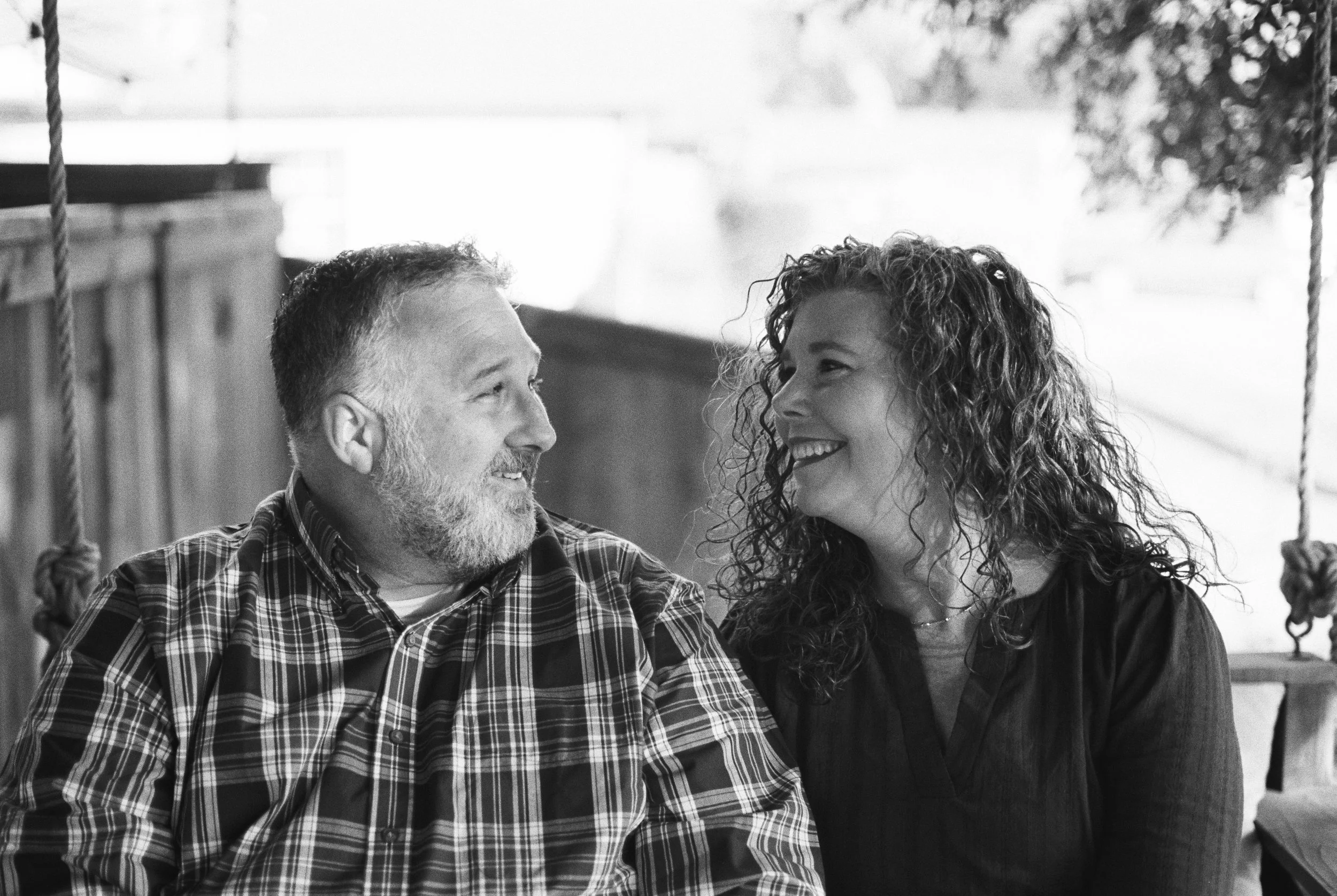A black and white photo of a man with a beard and a woman with curly hair, both smiling and looking at each other.