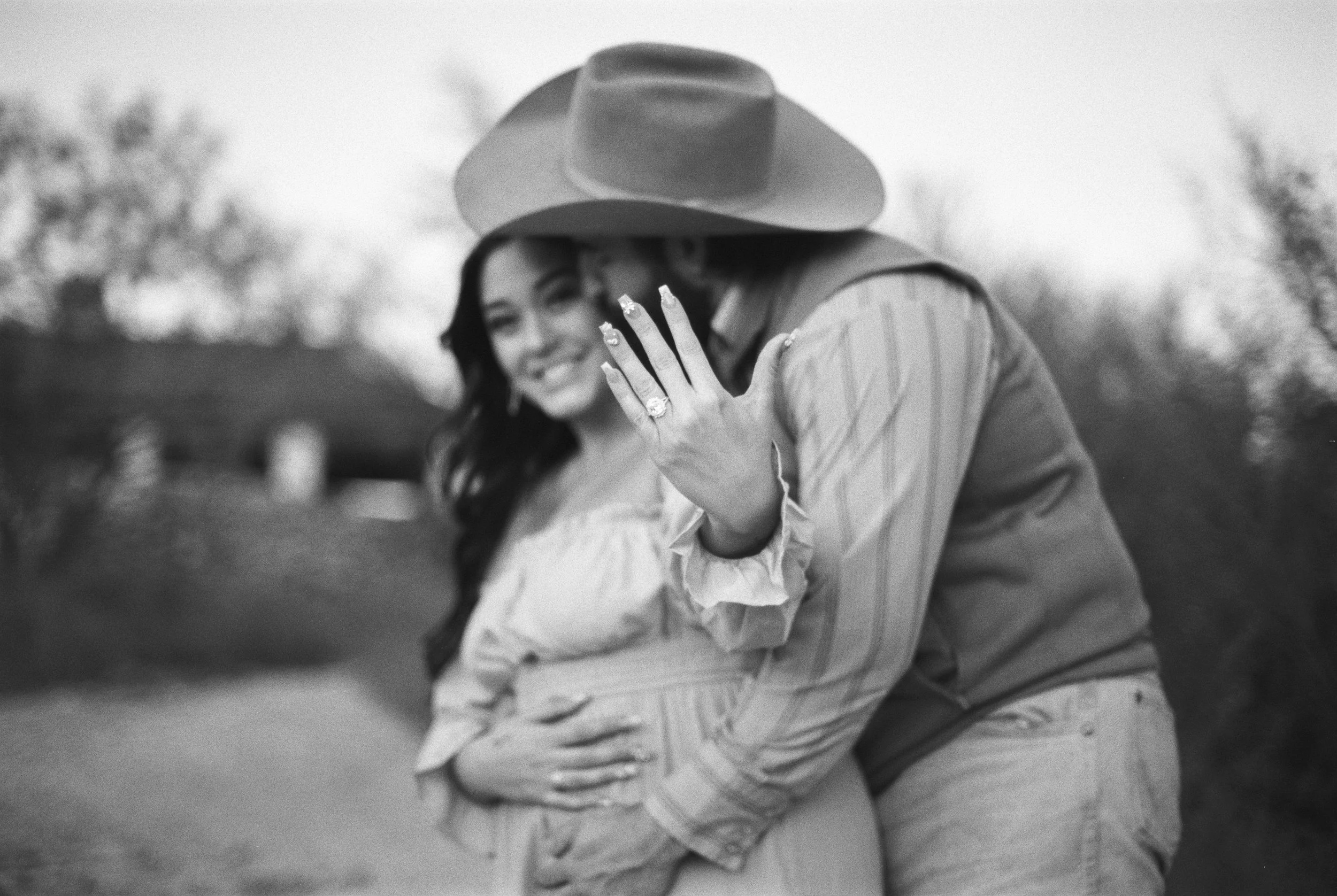 A woman showing off her engagement ring with a man embracing her from behind, outdoors in a natural setting.