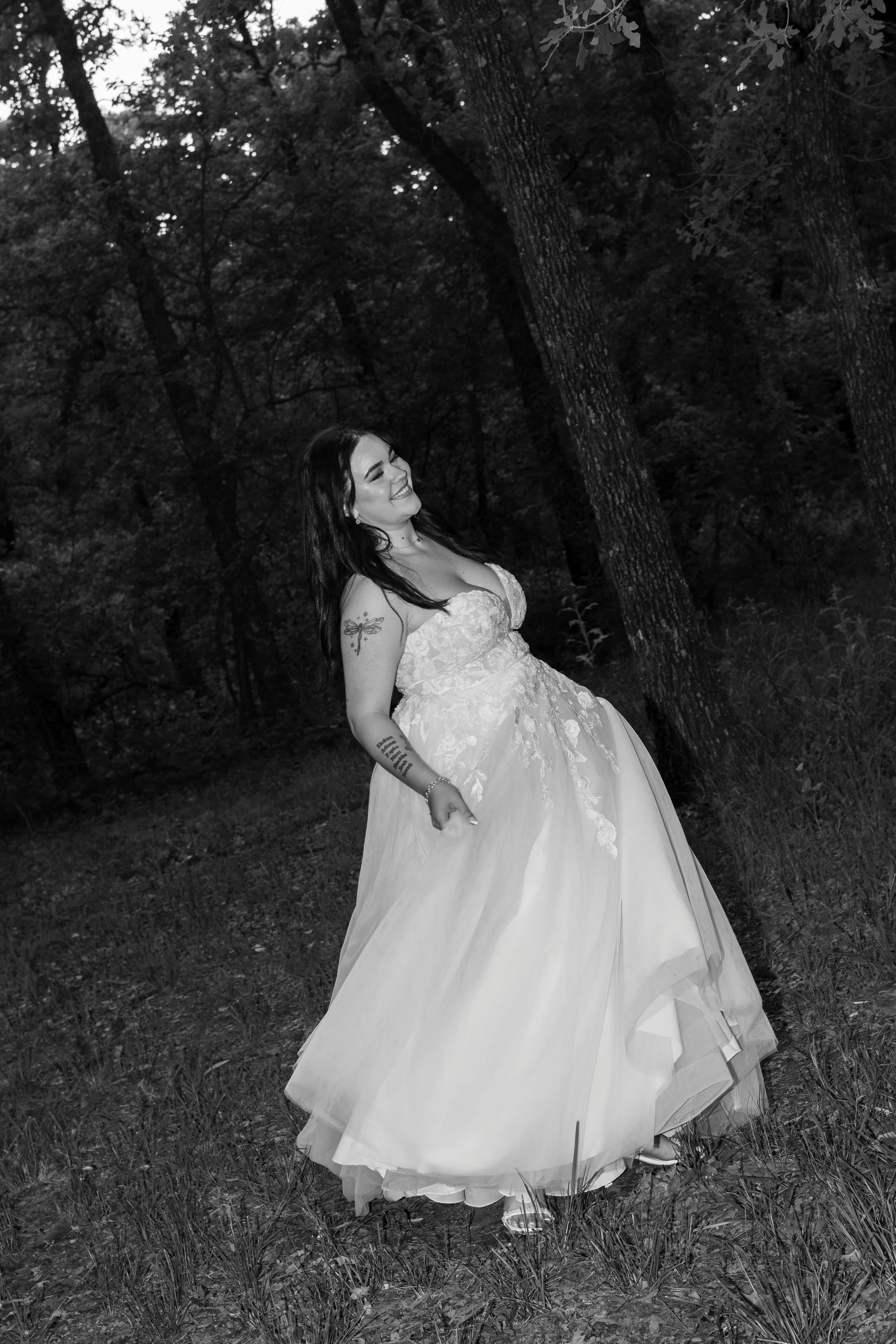 A woman in a wedding dress with a tattoo on her left arm, smiling and standing outdoors among trees and grass in a wooded area.
