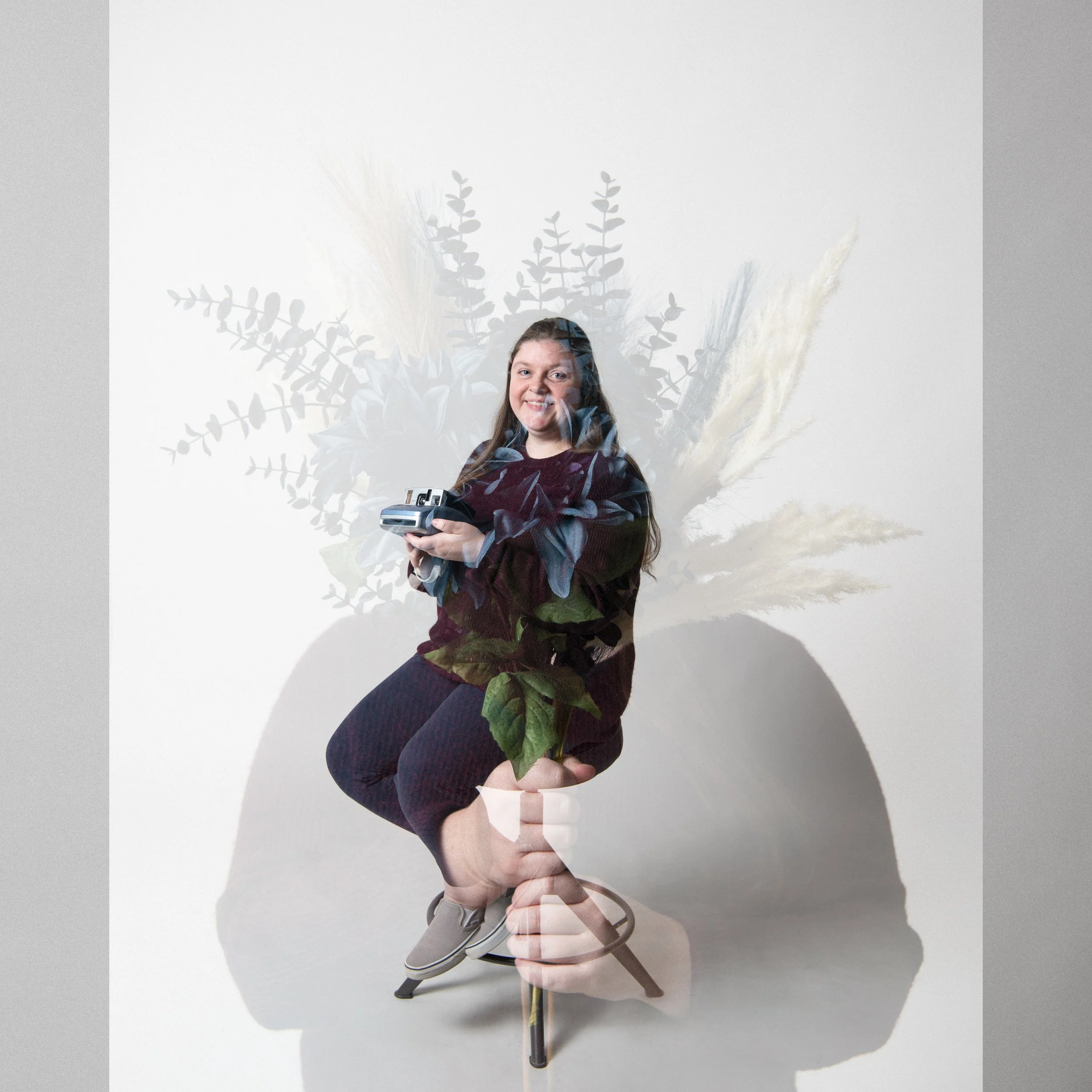 A woman sitting on a stool holding a camera, with a layered shadow of a plant behind her and an upside-down hand holding a small potted plant in the foreground.
