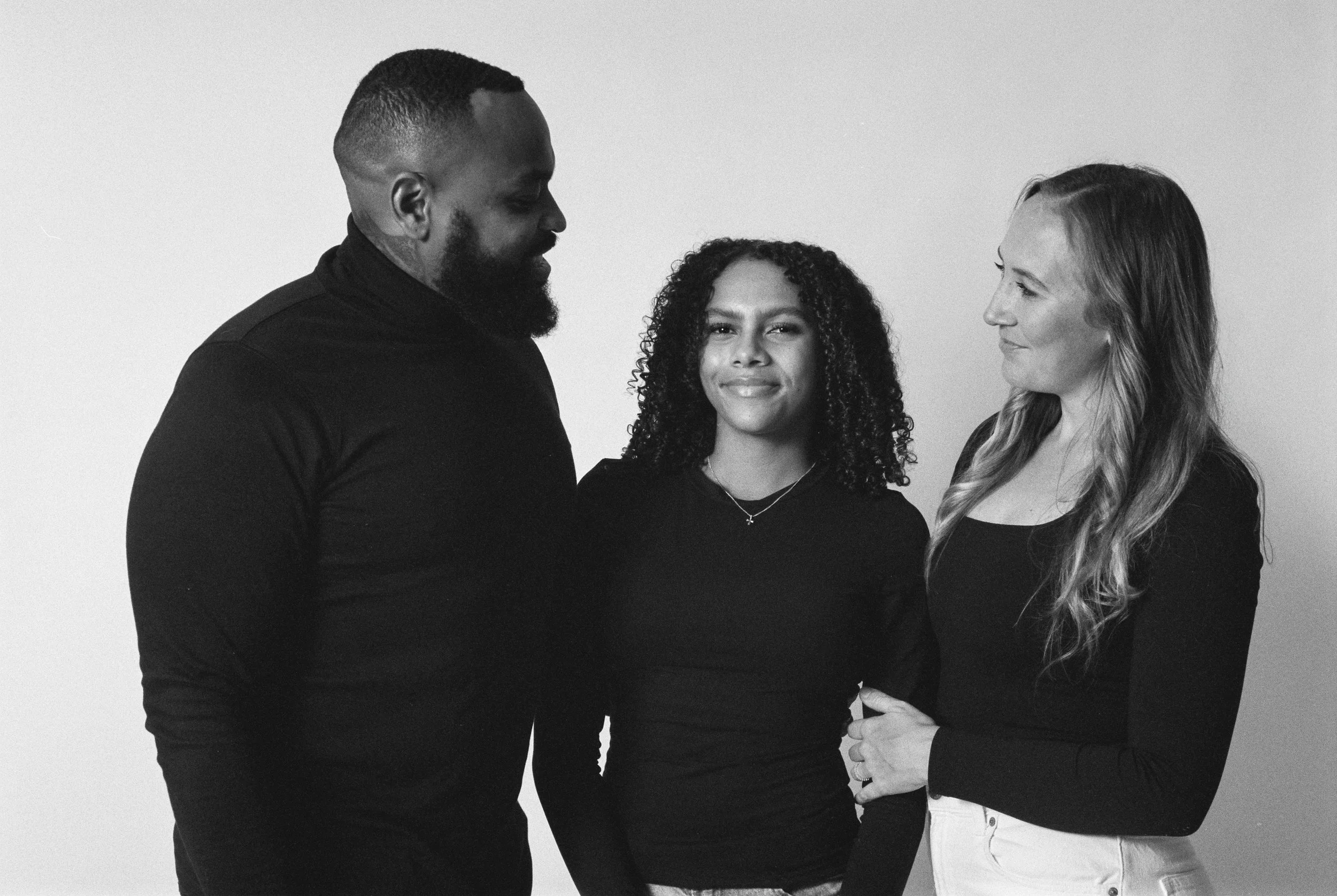 Black and white photo of three women standing close together, smiling and looking at each other, against a plain background.