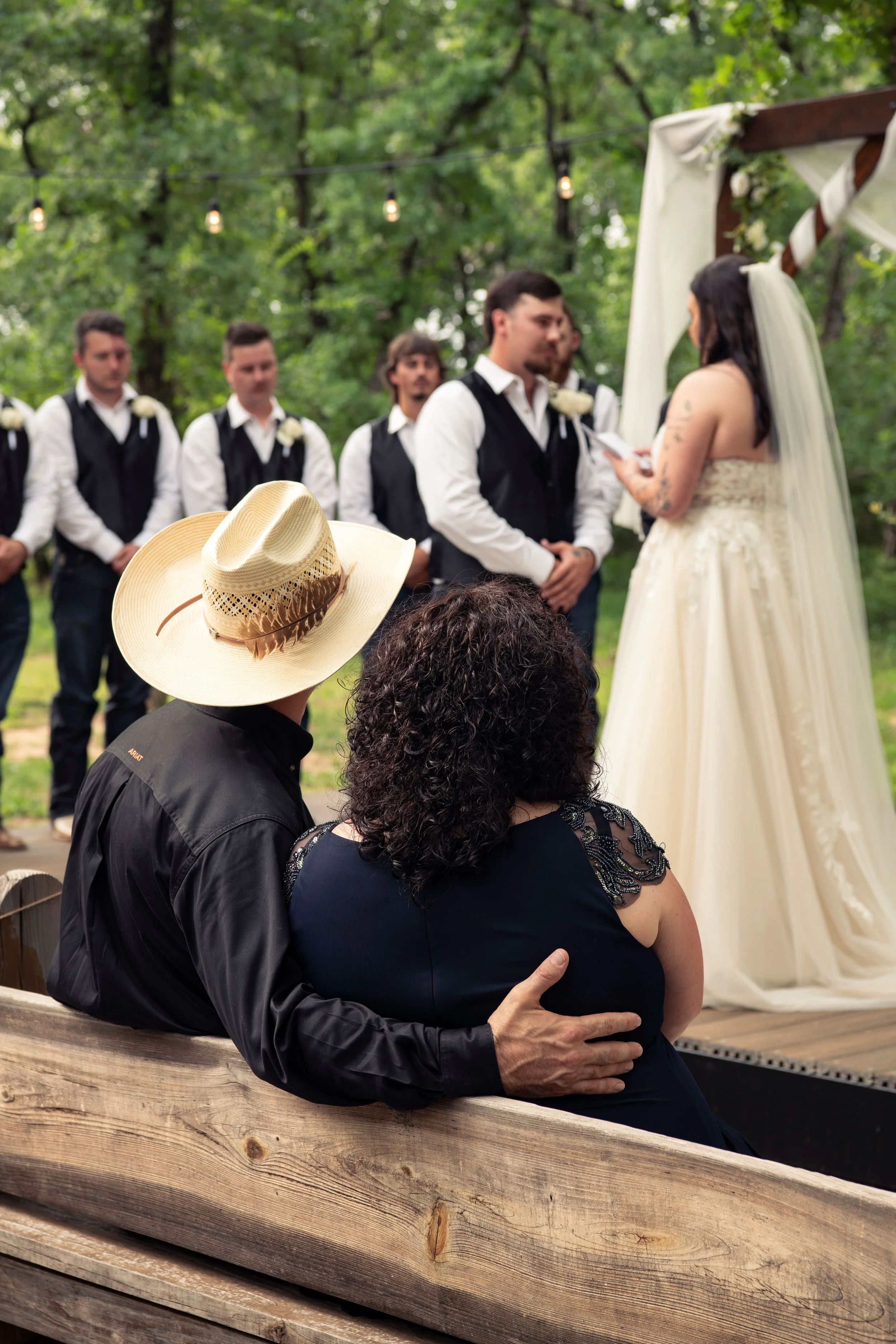 A couple is standing at a wedding altar outdoors surrounded by trees, exchanging vows. An officiant is holding a paper, seemingly reading the vows. Bridesmaids and groomsmen stand in the background. In the foreground, a guest wearing a straw hat and 