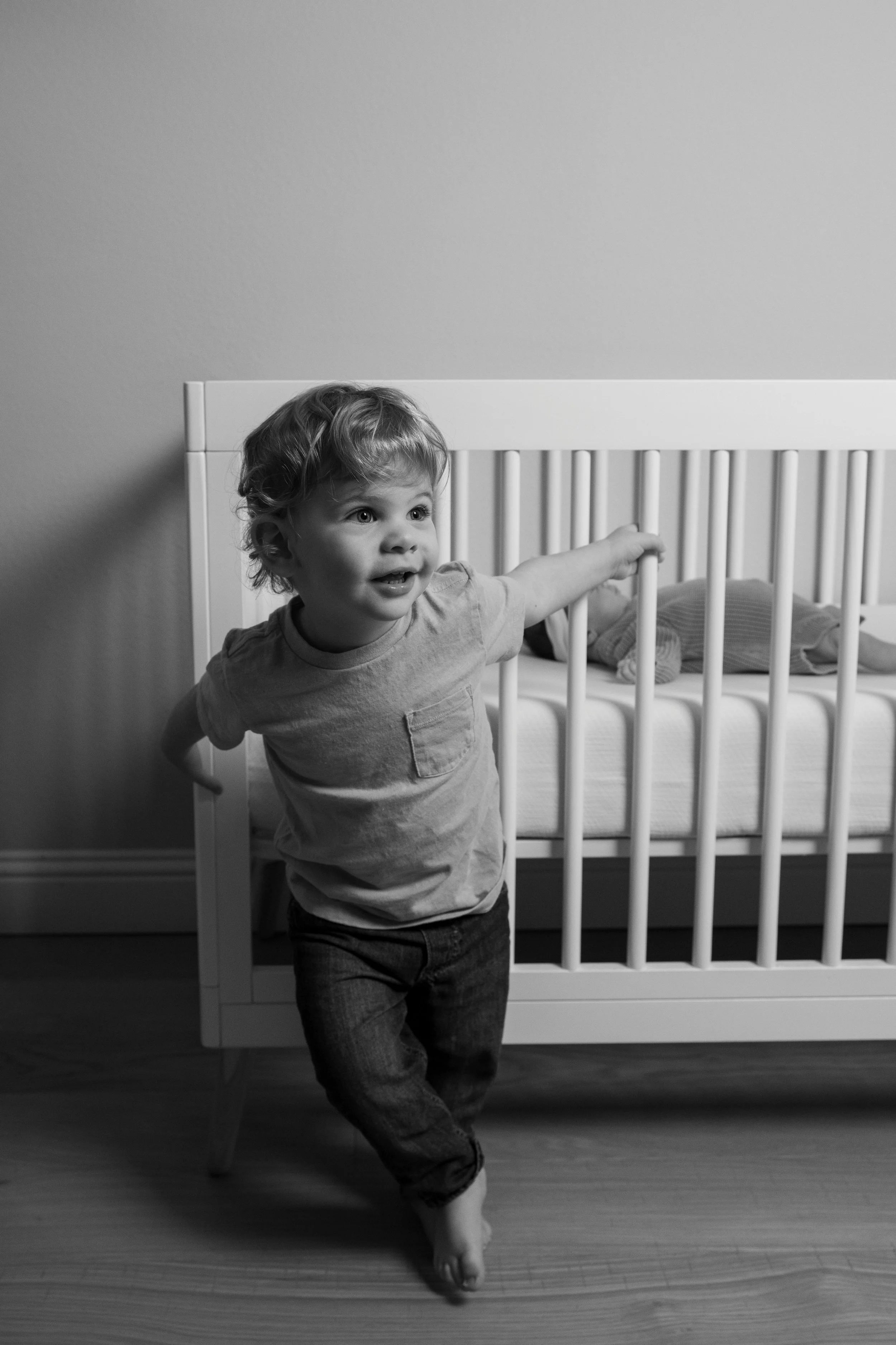 A young child with curly hair stands next to a crib, smiling and reaching out with one arm, in a room with a plain wall.