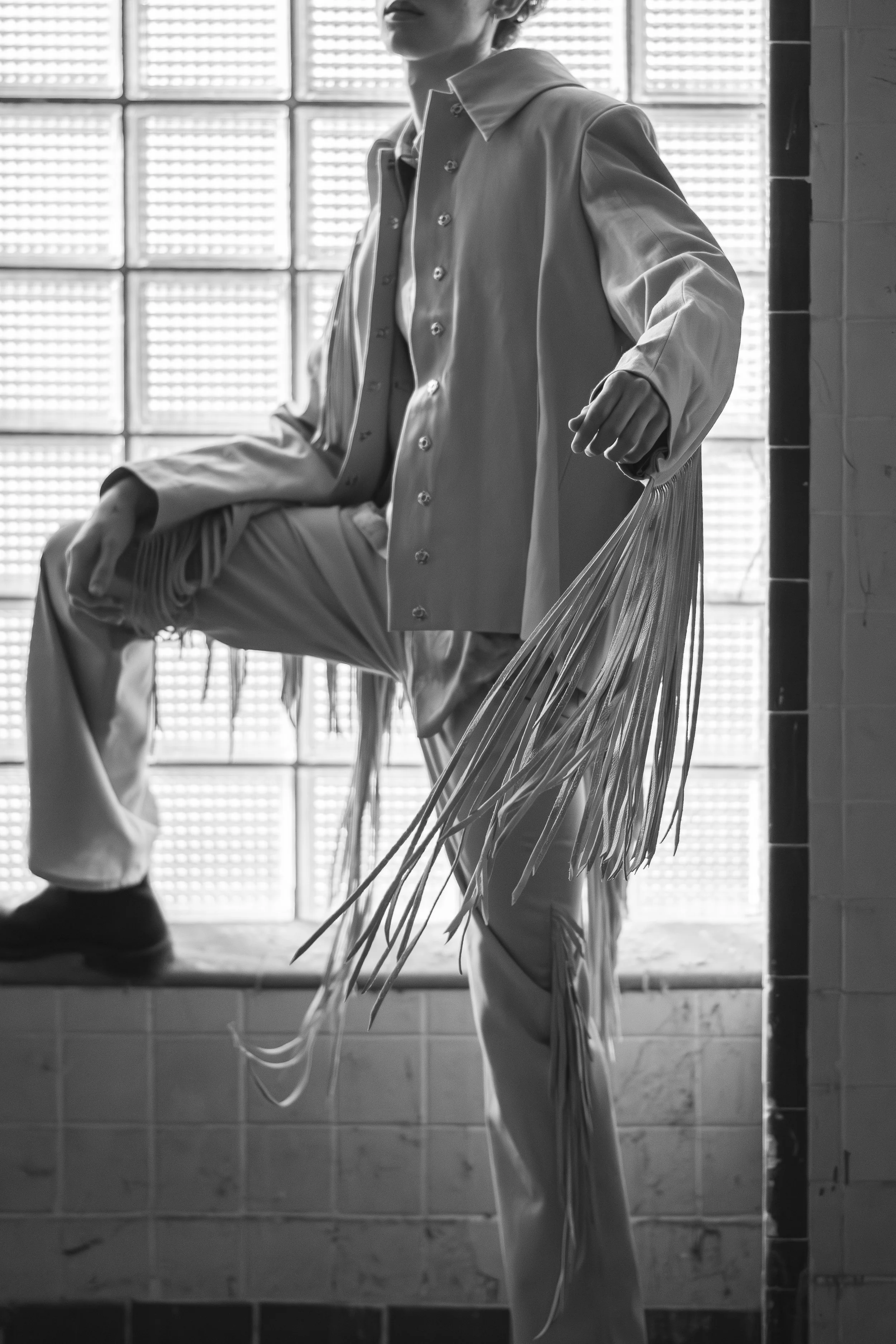 Black and white photo of a person sitting with one leg crossed, wearing a long coat with fringe details on the sides, in front of a glass block wall, with a partially visible face and holding a plastic bag.