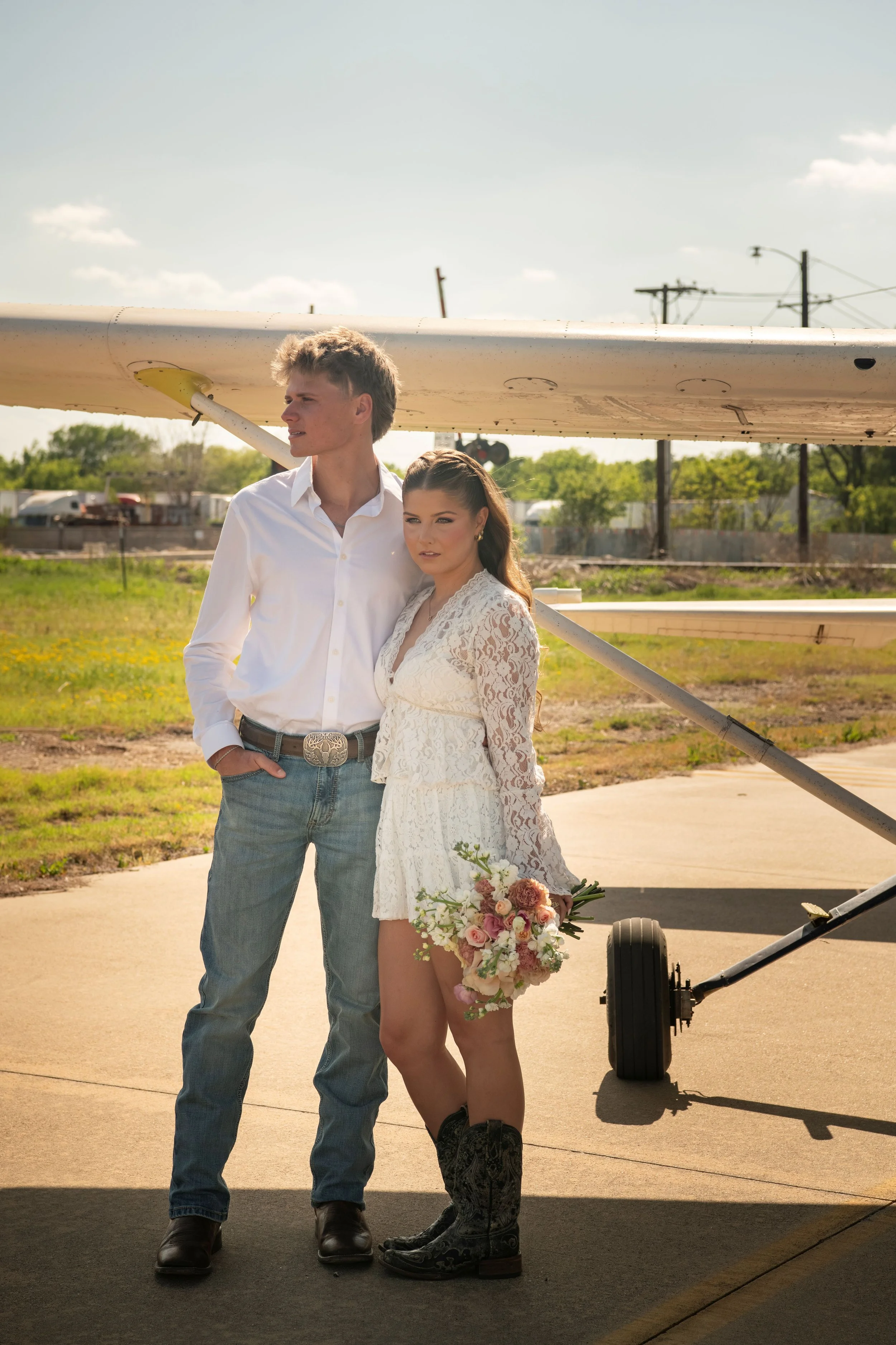 A young man and woman stand outdoors near a small airplane, with the woman holding a bouquet of flowers, both dressed in white lace outfits, and the man wearing blue jeans and the woman in cowboy boots.