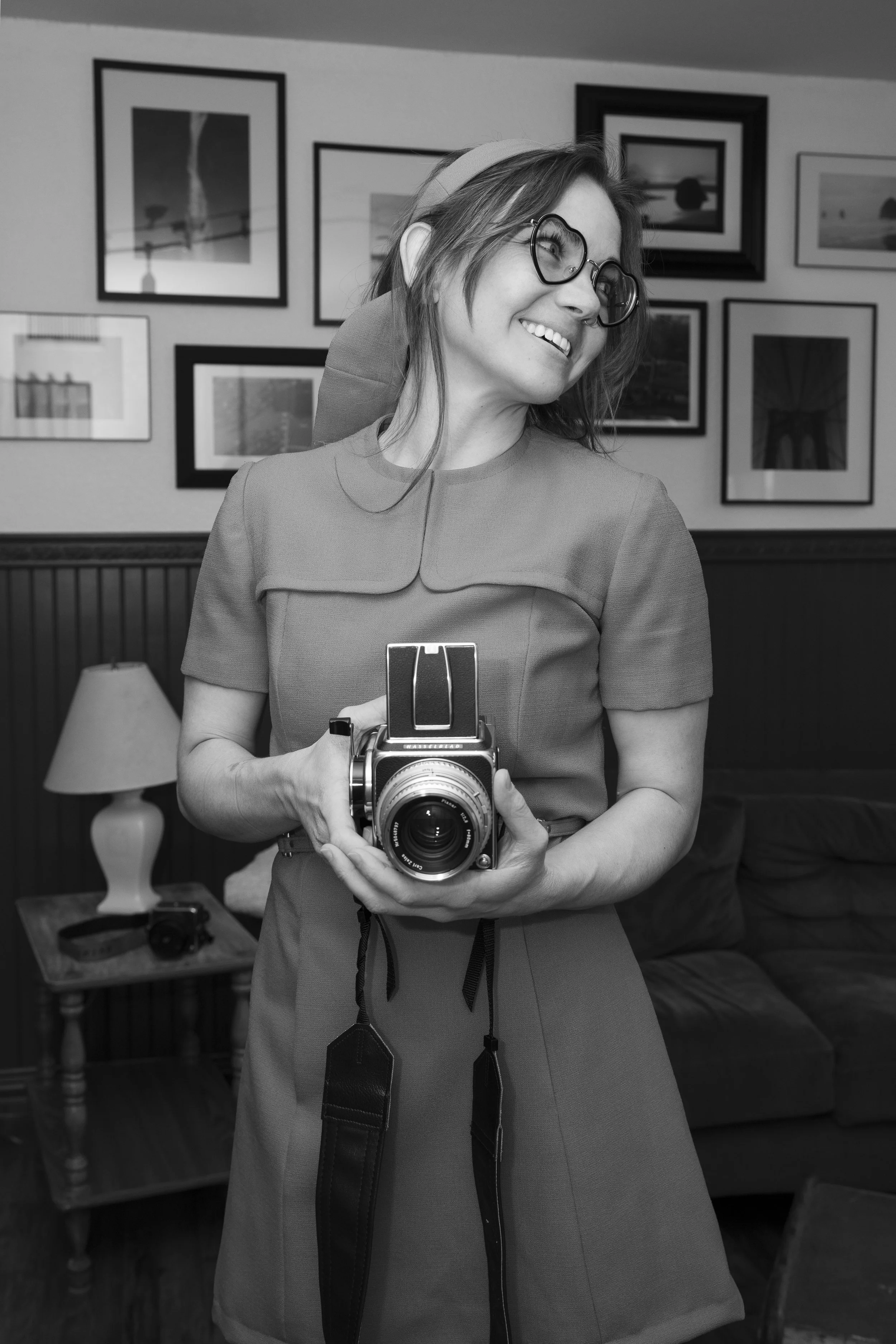 A woman with glasses and a vintage dress, holding a camera, smiling in a room with framed pictures on the wall.