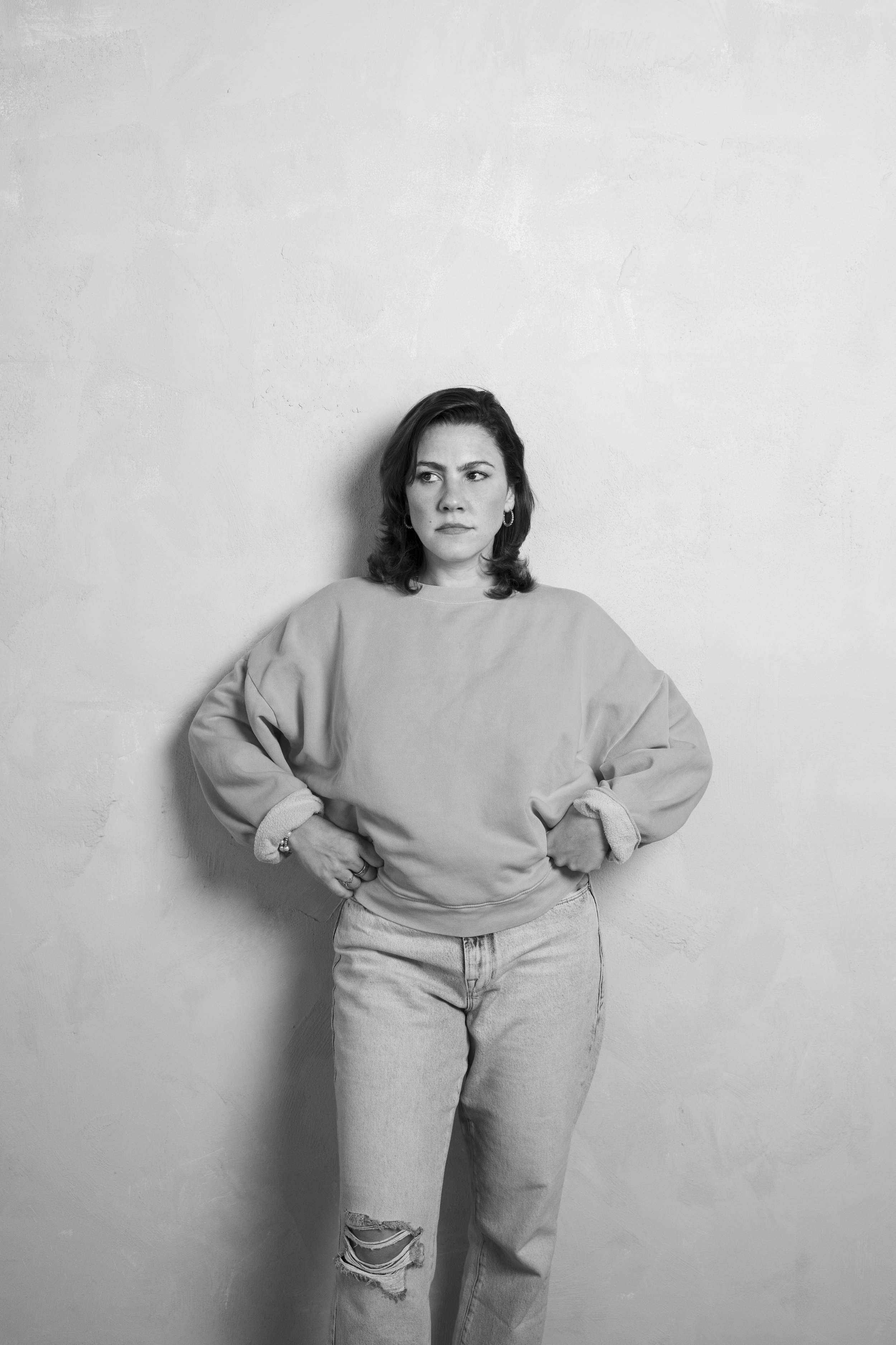 Black and white photo of a woman standing against a plain wall, wearing a casual sweatshirt and ripped jeans, with a serious expression.