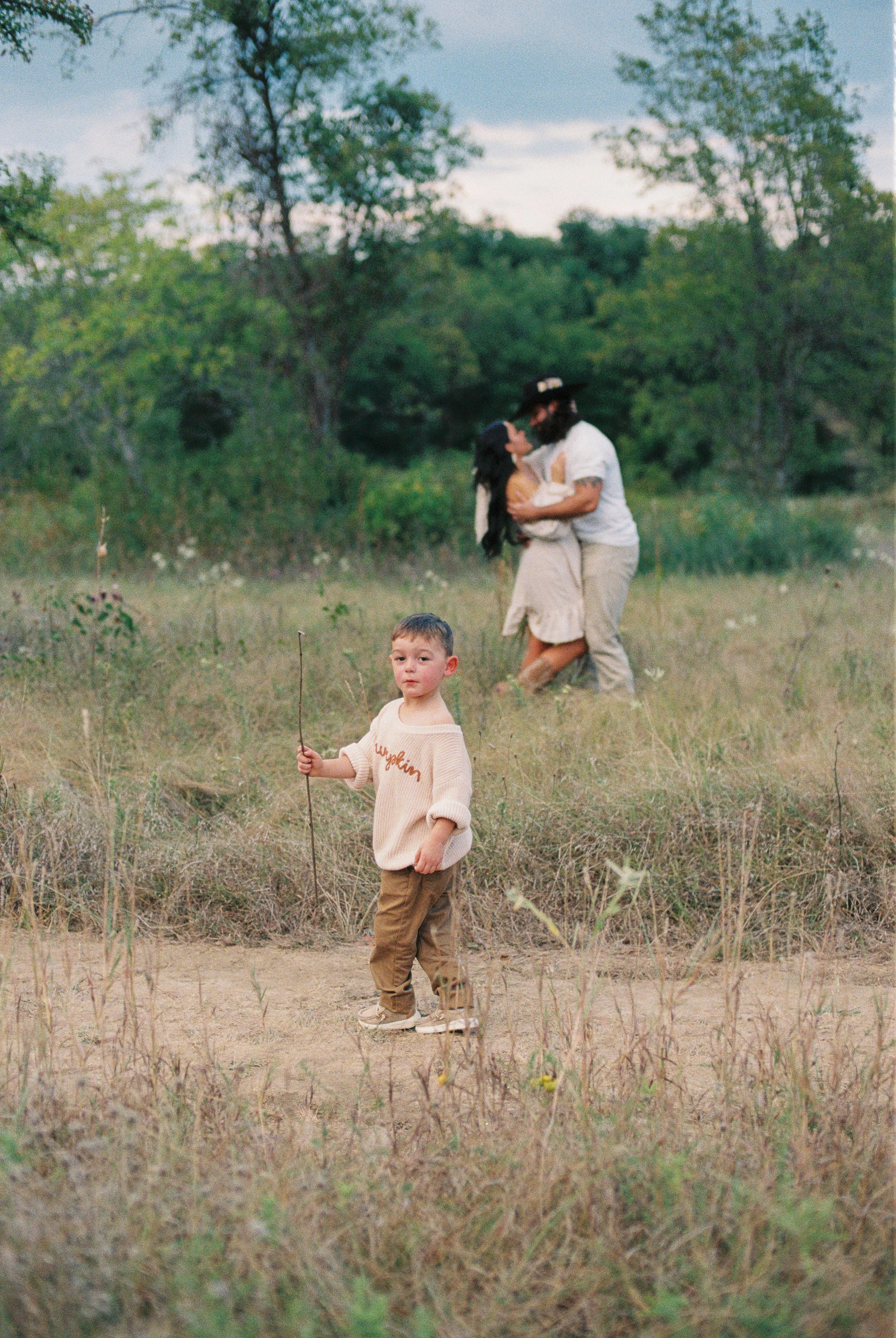 A young boy standing on a dirt path holding a stick, with a couple kissing in the background on a grassy field surrounded by trees.