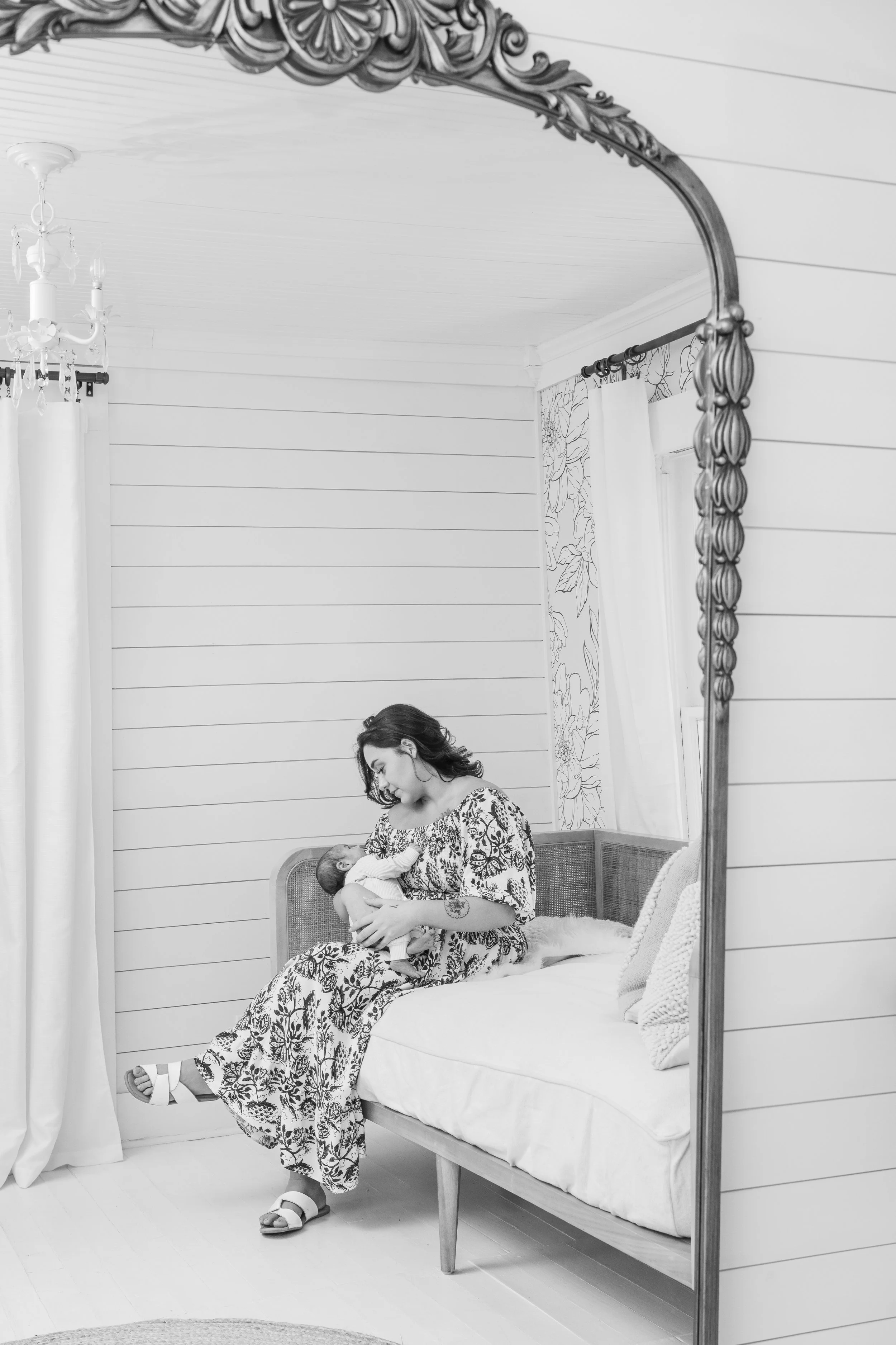 A woman in a patterned dress sitting on a bench, breastfeeding a baby, with her head slightly bent toward the child, reflected in a large ornate mirror in a bright, cozy room.