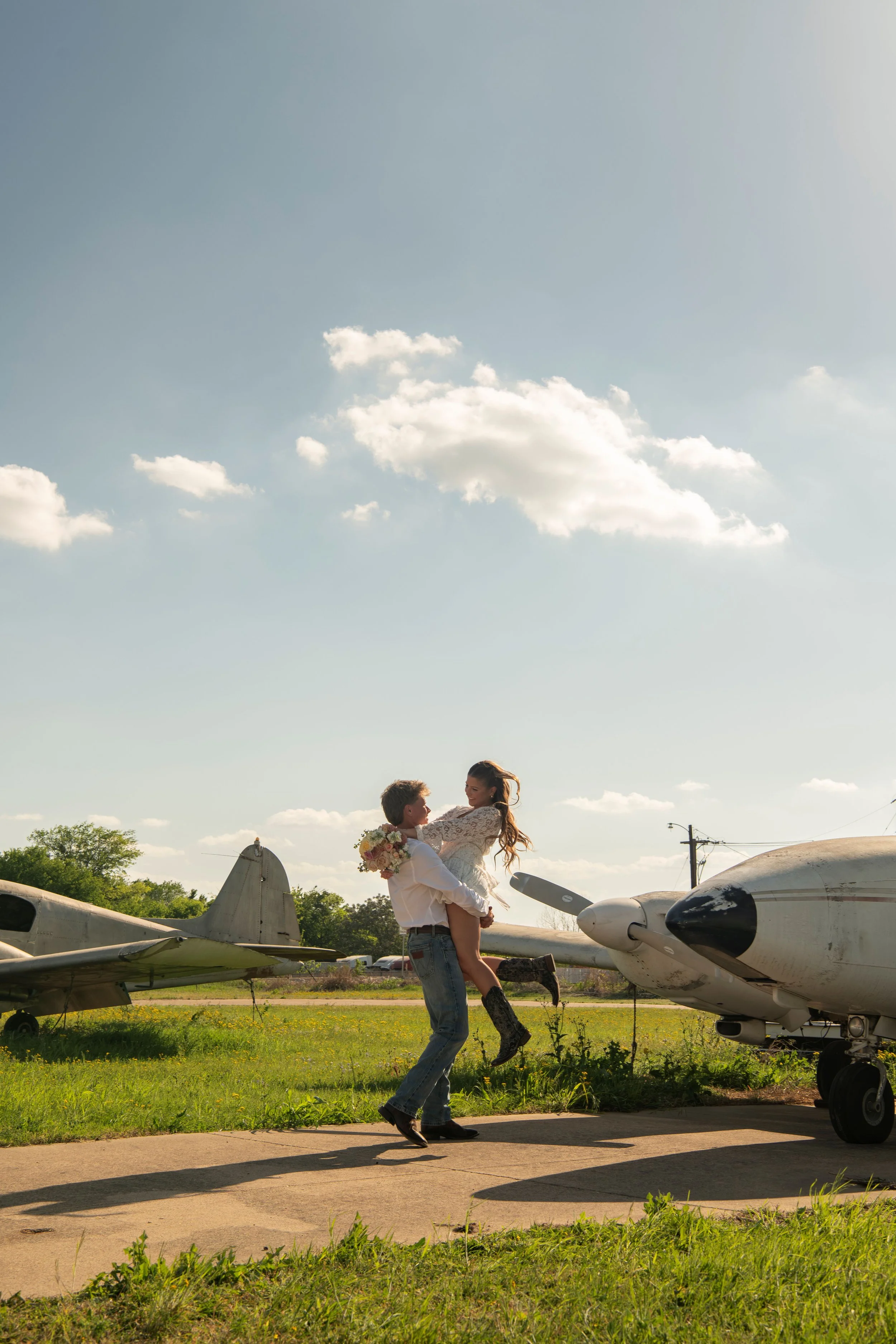 A man lifting a woman in a white dress holding a bouquet of flowers at an airfield with old airplanes and airplanes in the background under a partly cloudy sky.