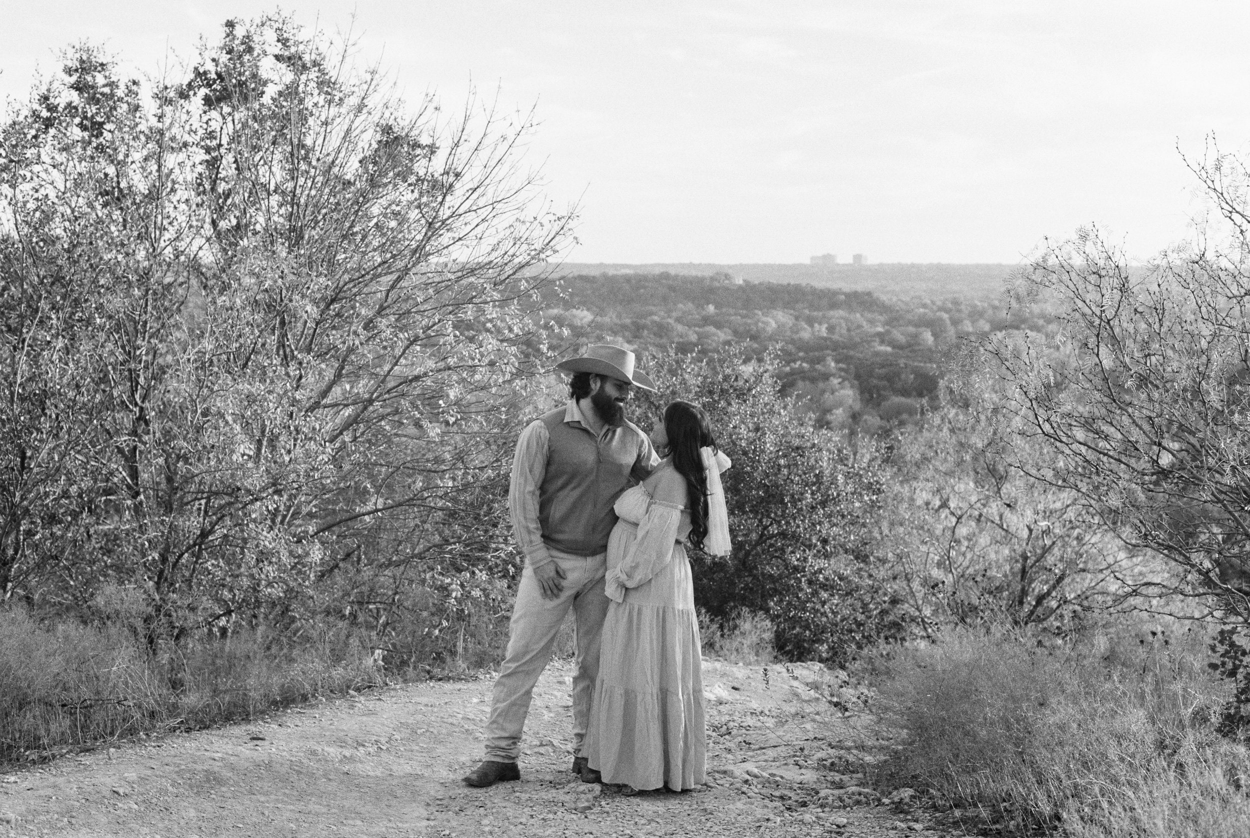 Black and white photo of a man and woman standing on a dirt path outdoors, holding hands, surrounded by trees and bushes with a distant landscape and buildings in the background.