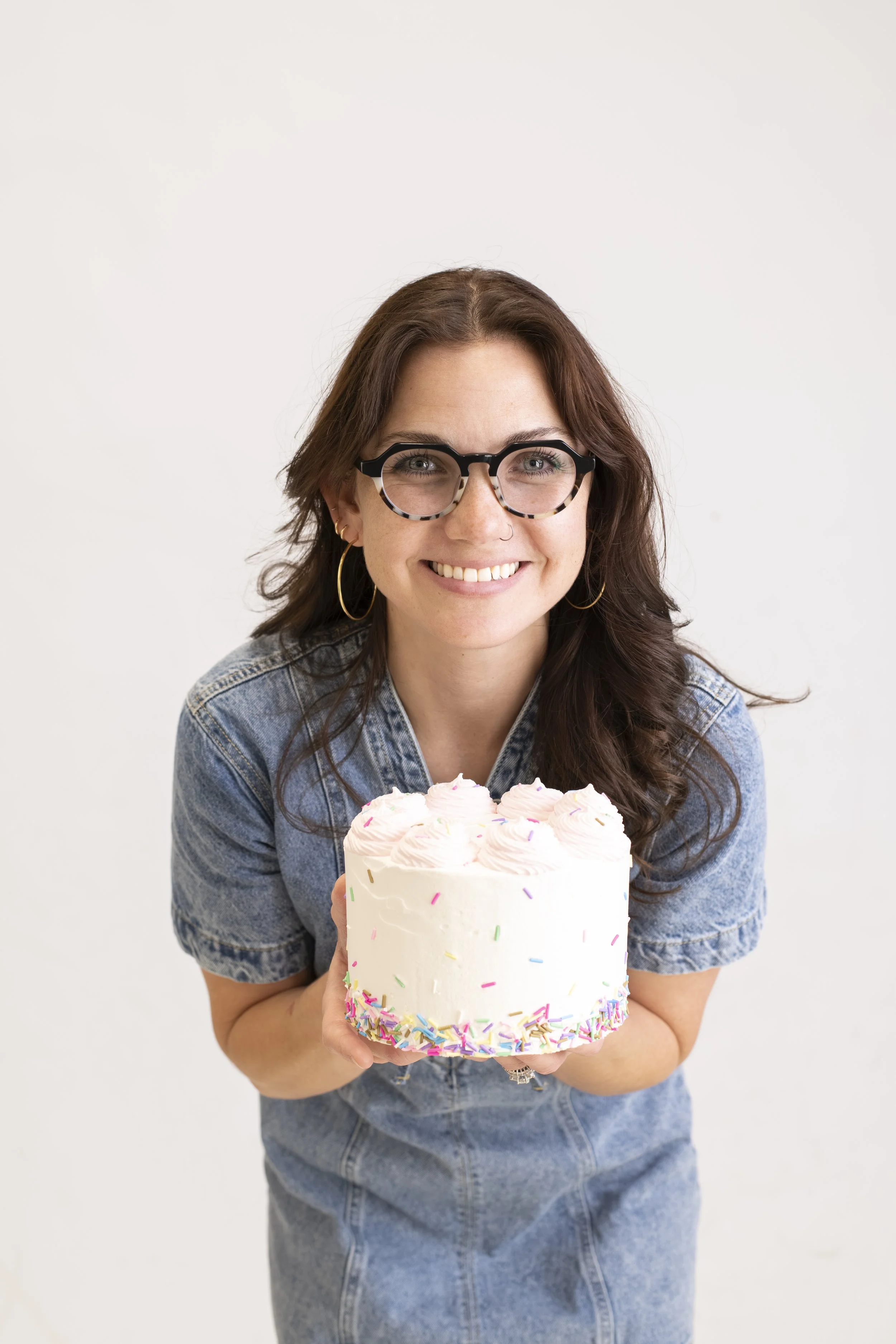 A woman with glasses and hoop earrings holding a birthday cake decorated with pink frosting and rainbow sprinkles, smiling at the camera.