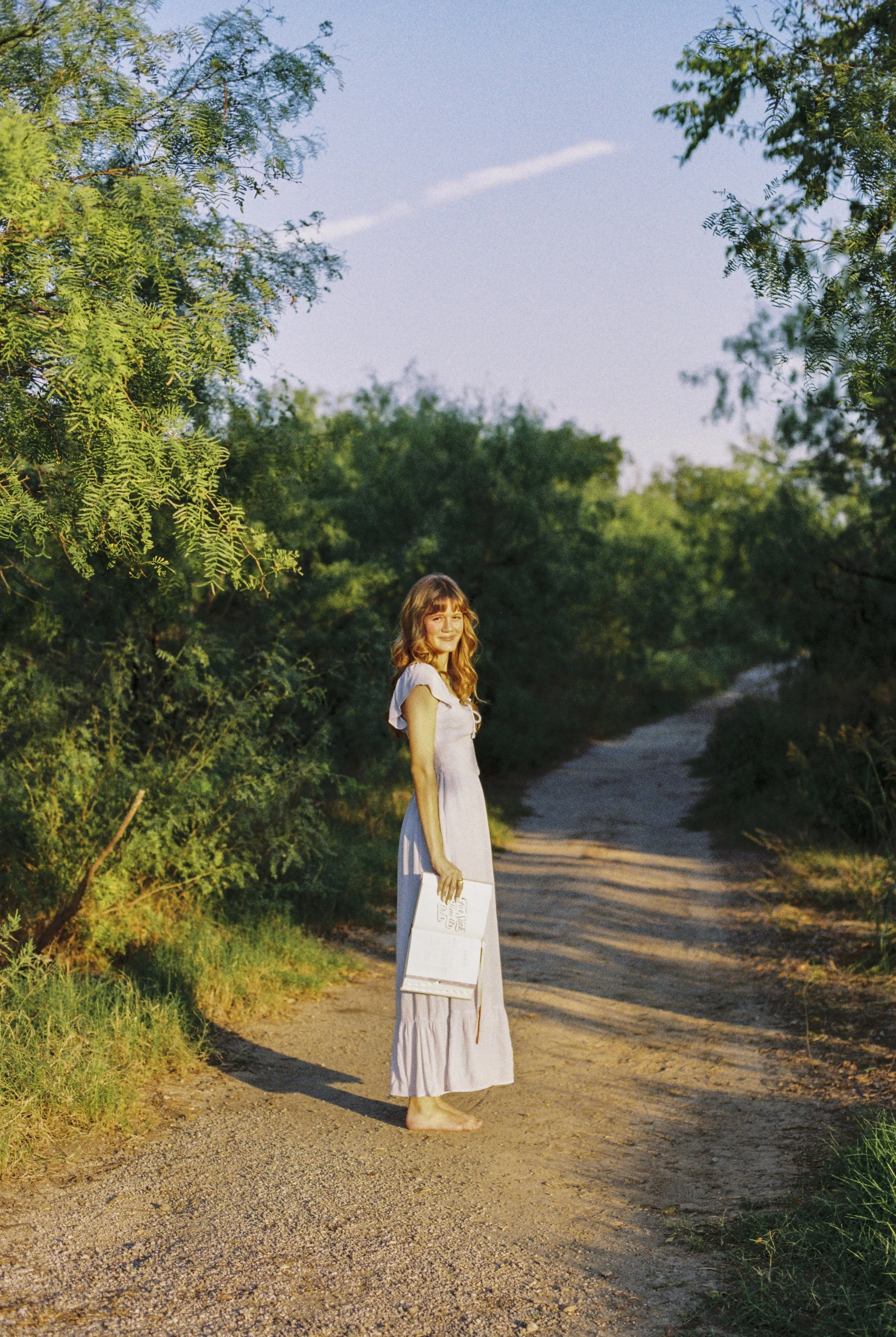 A woman with long, wavy hair wearing a light-colored dress, barefoot, standing on a dirt trail surrounded by green trees and bushes, holding a notebook or sketchpad in one hand, during the daytime.