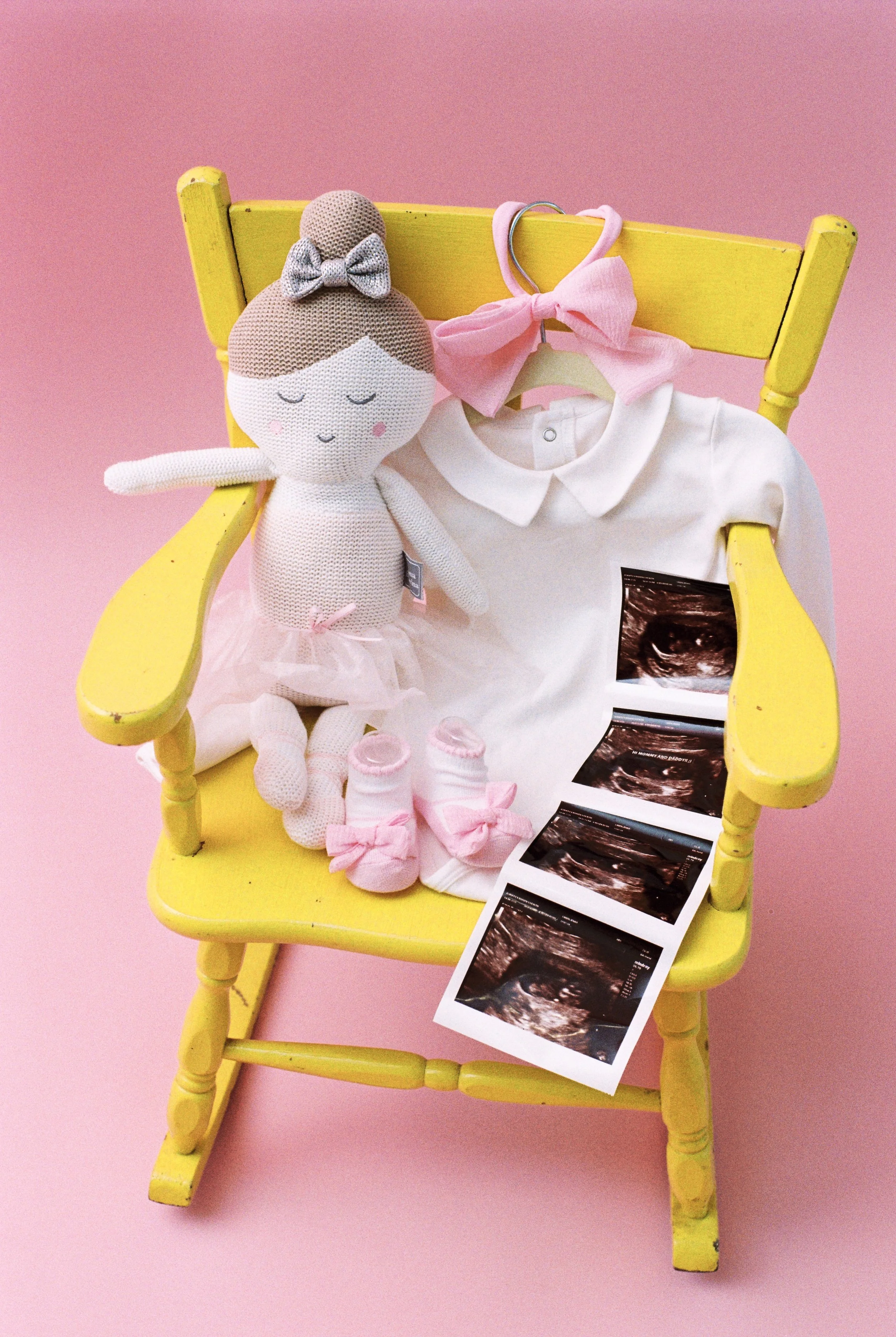 A yellow rocking chair against a pink background displays a crochet doll, baby clothes, and ultrasound images, indicating a pregnancy announcement.