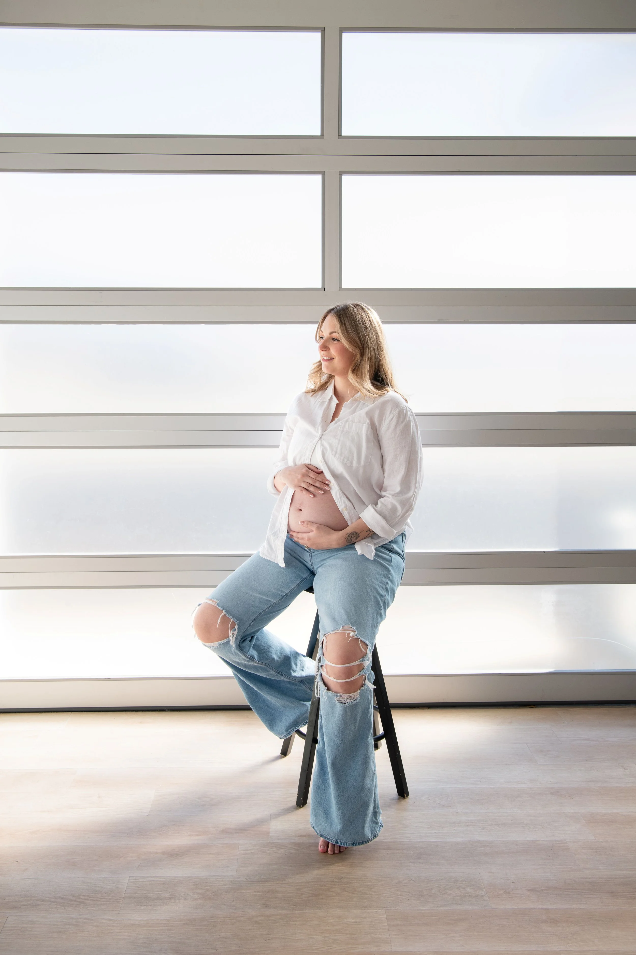 A pregnant woman sitting on a stool, smiling, with her hand on her belly, in a bright room with large windows.