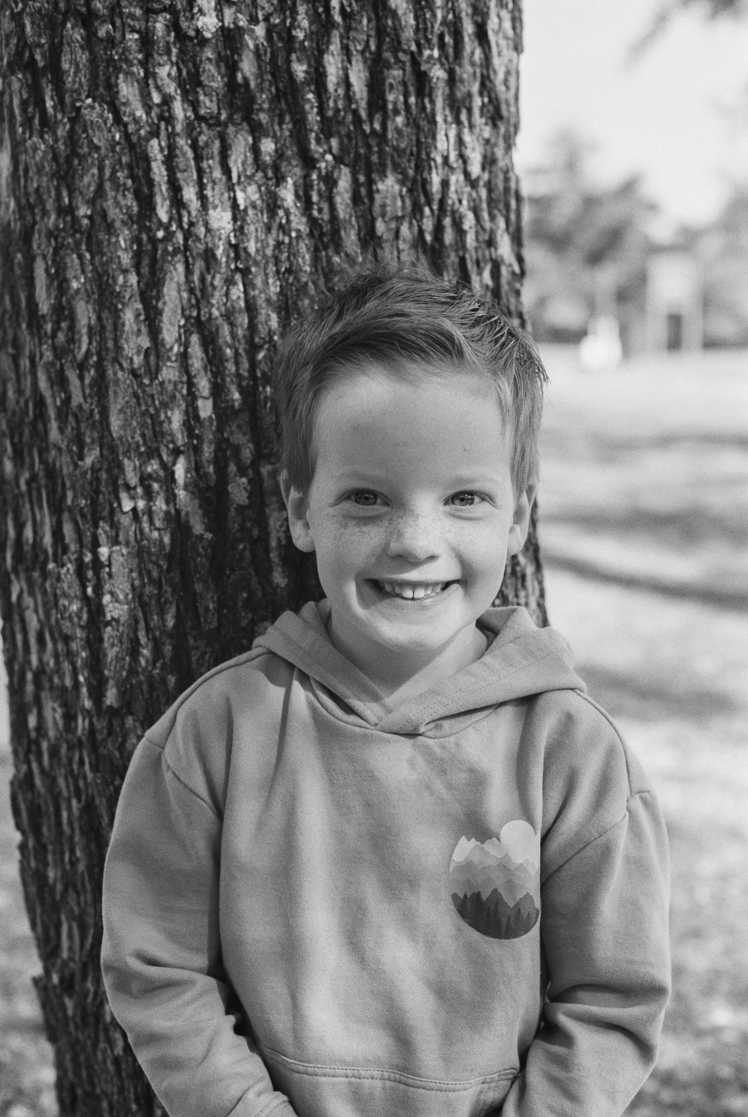 A young boy with short hair and freckles, smiling and wearing a hoodie, leaning against a tree outdoors.