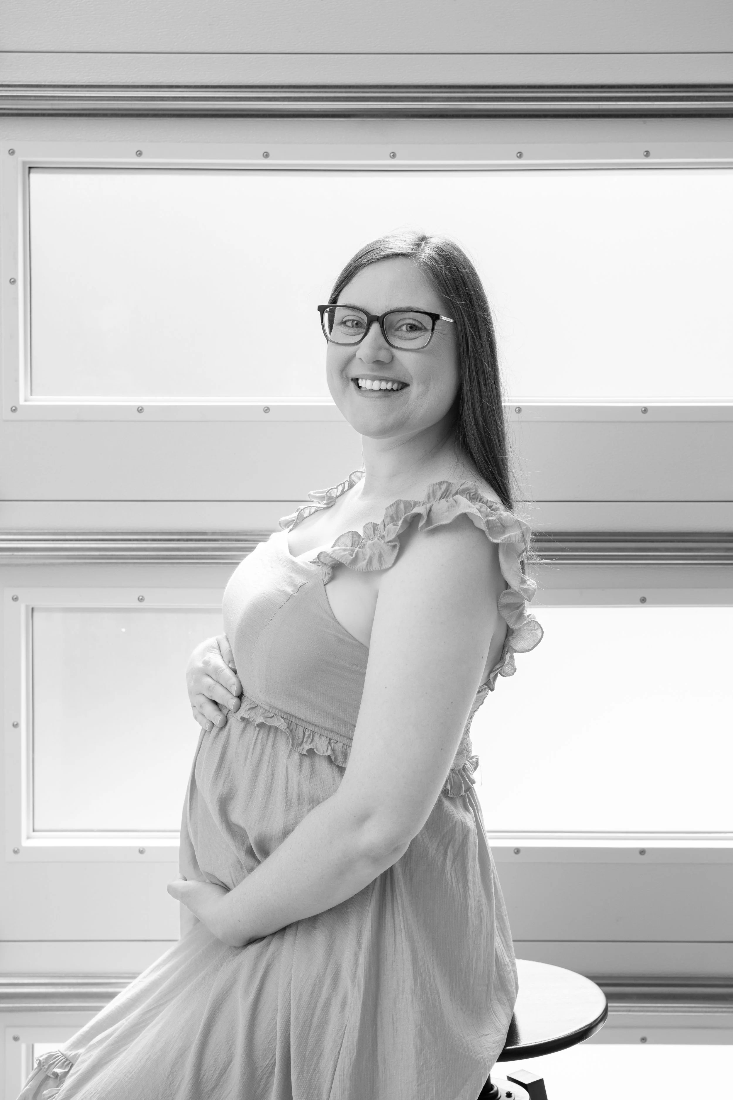 A smiling woman with glasses sitting on a stool in front of a wall with rectangular windows, wearing a ruffled dress.