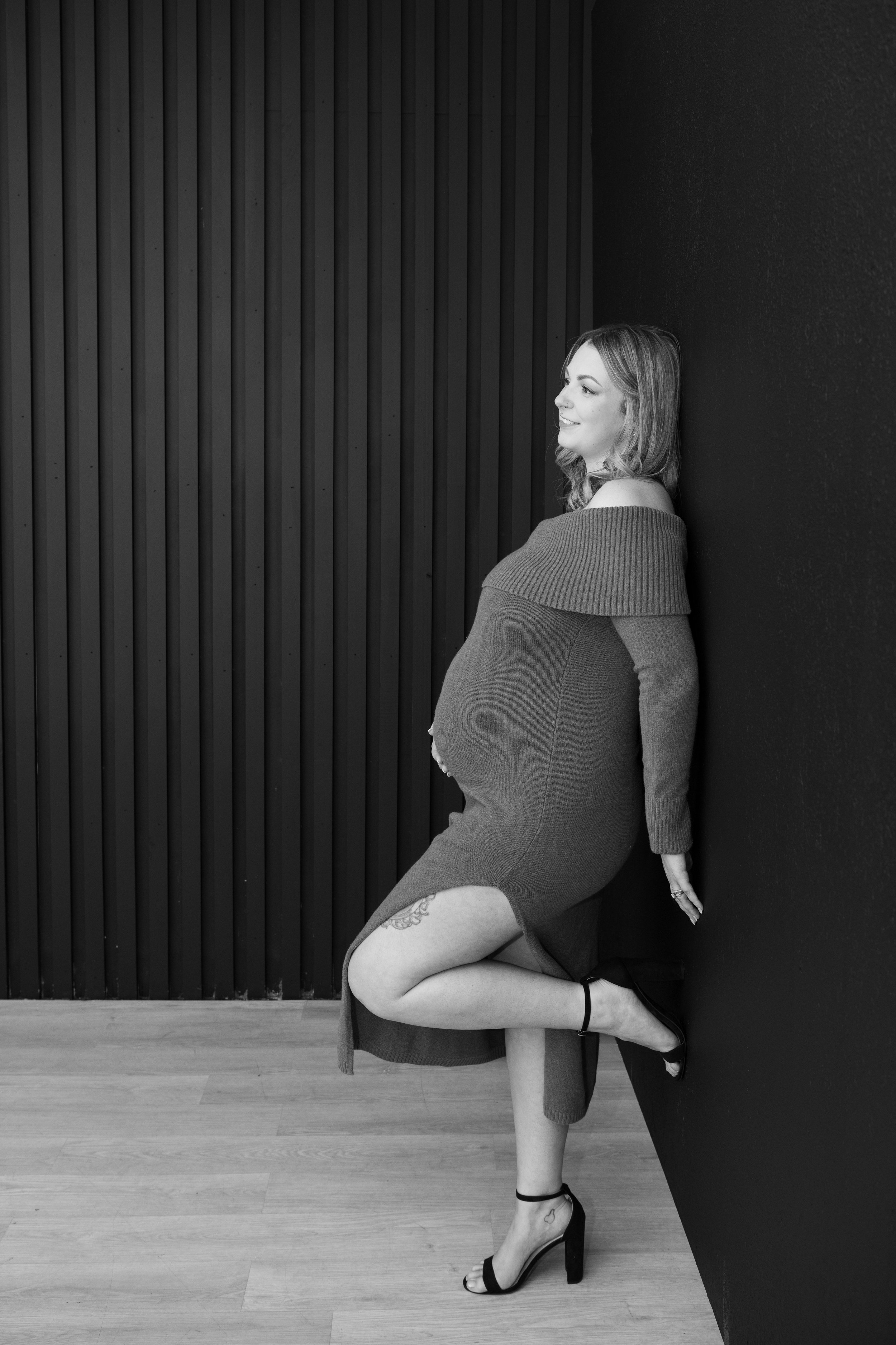 A pregnant woman in a long sweater dress, with one leg lifted and her foot against the wall, smiling and looking to the side, black and white photo.