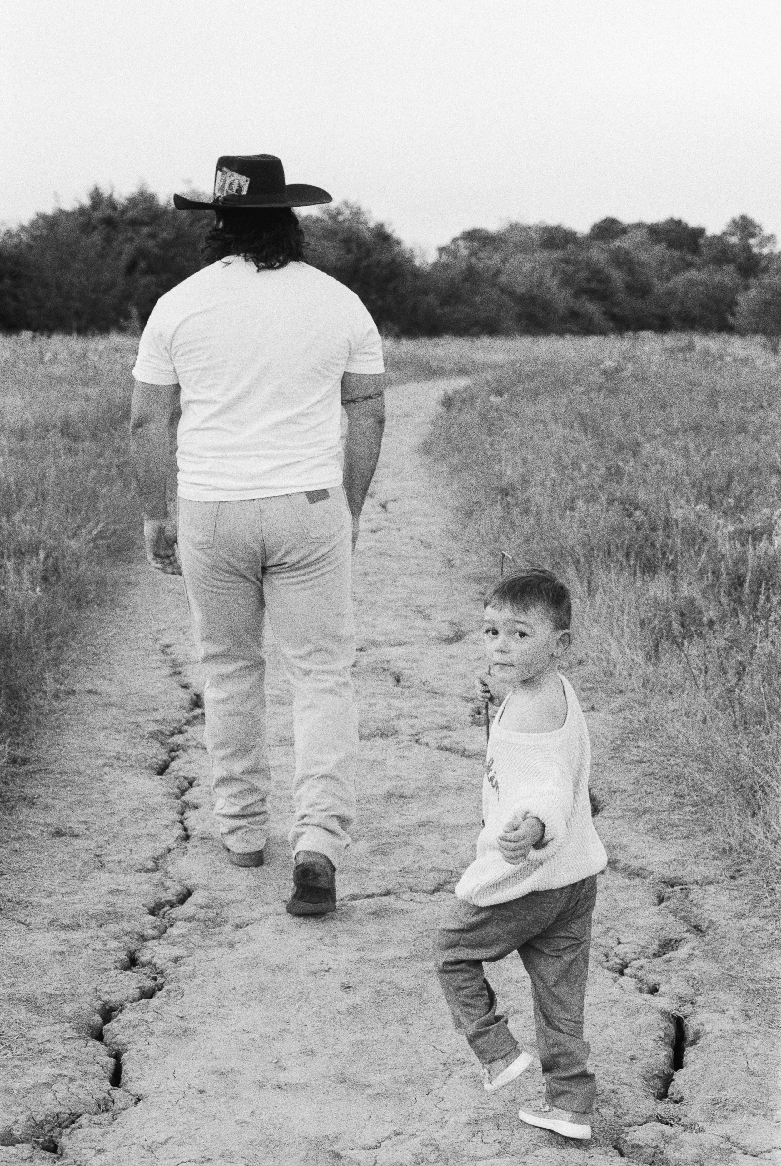 A man and a young boy walking on a cracked dirt path through a grassy field, with trees in the background, in black and white.
