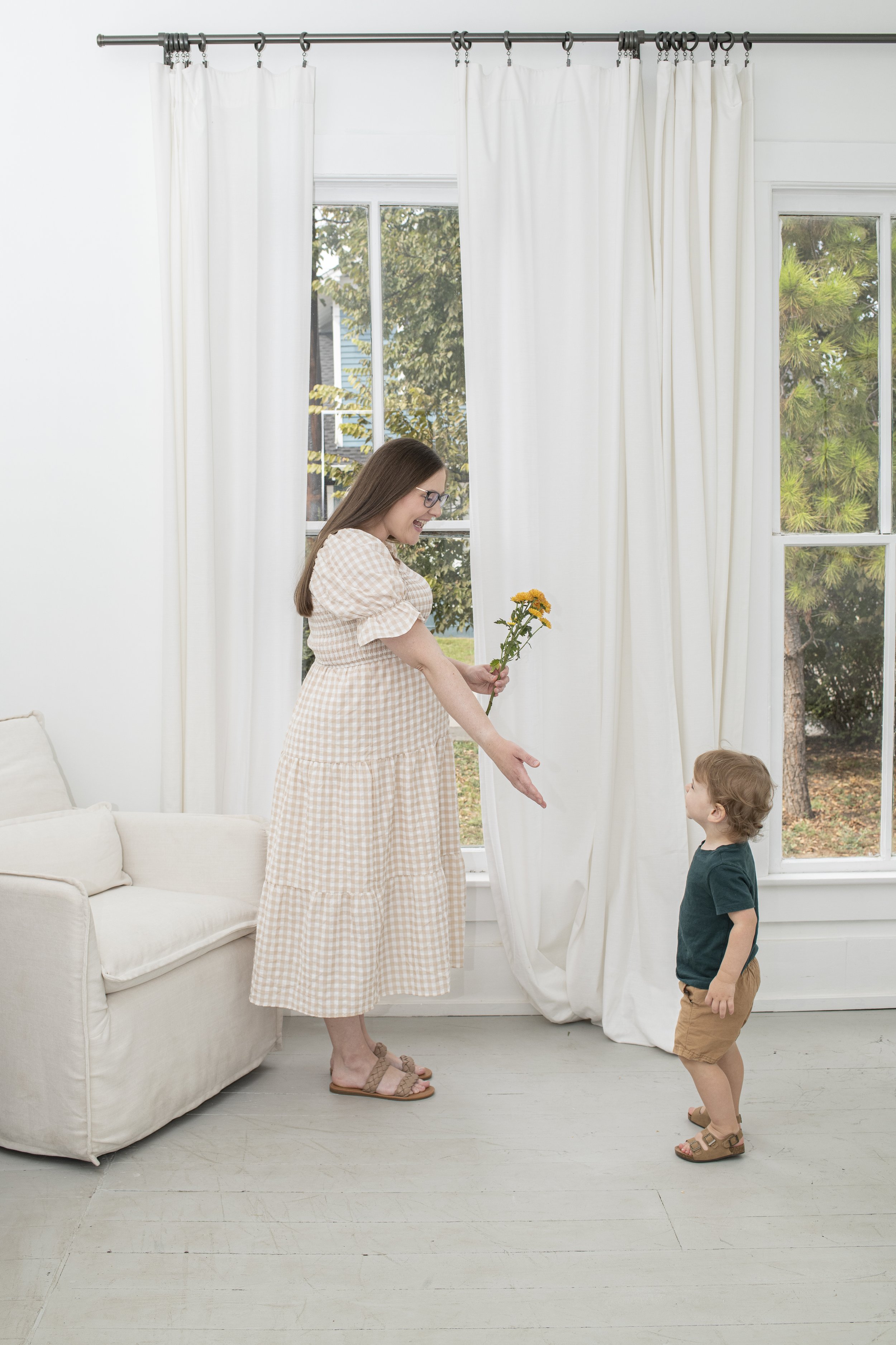 A woman giving a flower to a young boy in a bright room with white walls, large windows, and white curtains.
