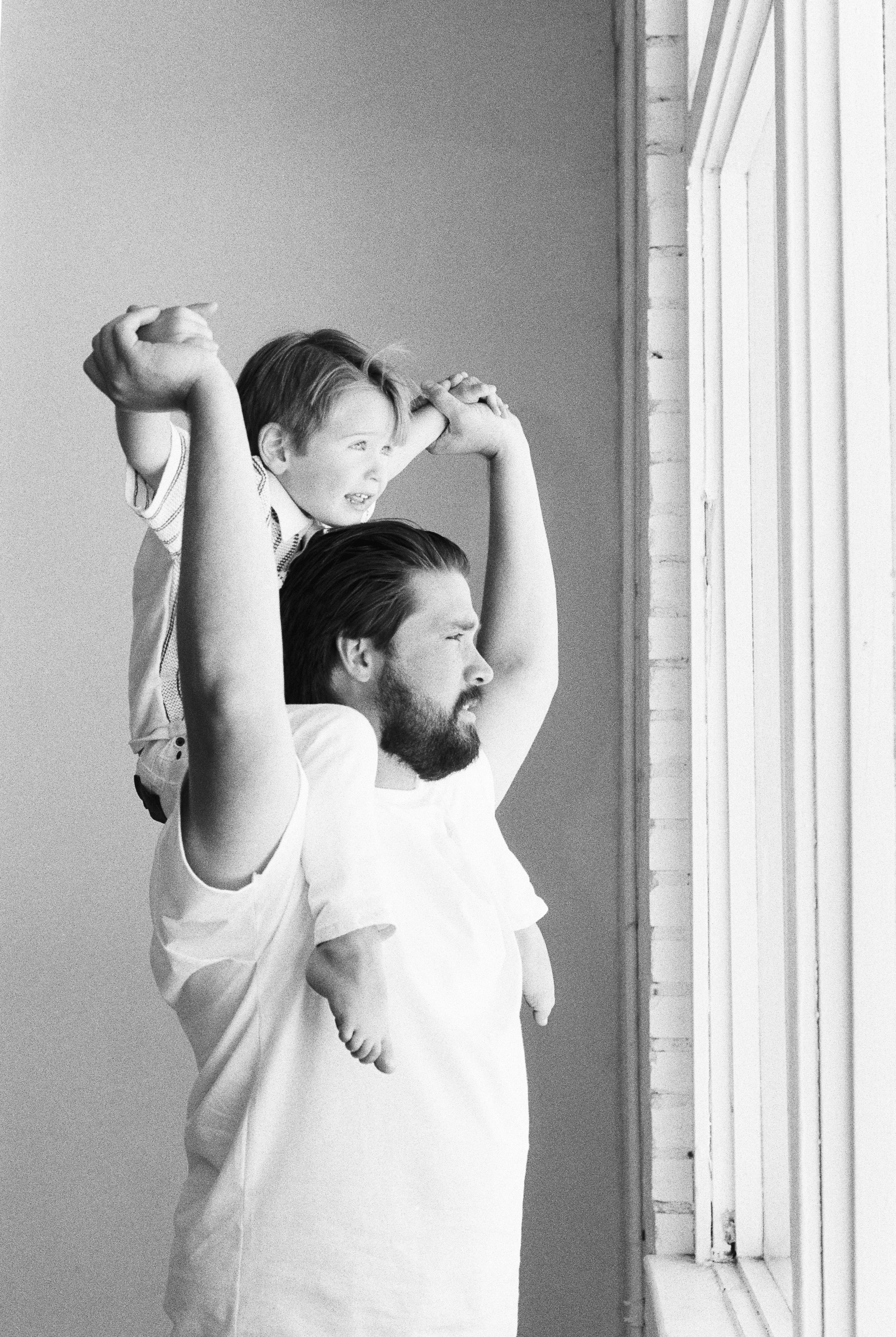 A man with a beard and a child sitting on his shoulders, looking out a window.