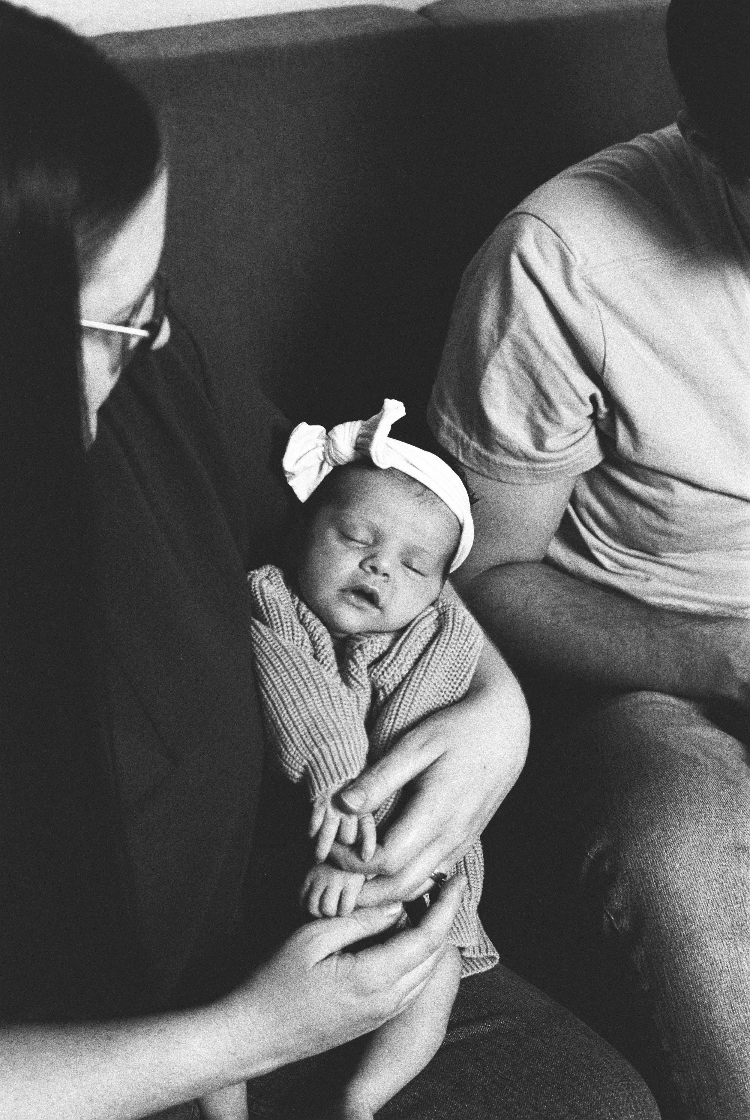 A sleeping baby girl with a bow on her head sitting on a woman's lap, surrounded by two adults, in black and white.