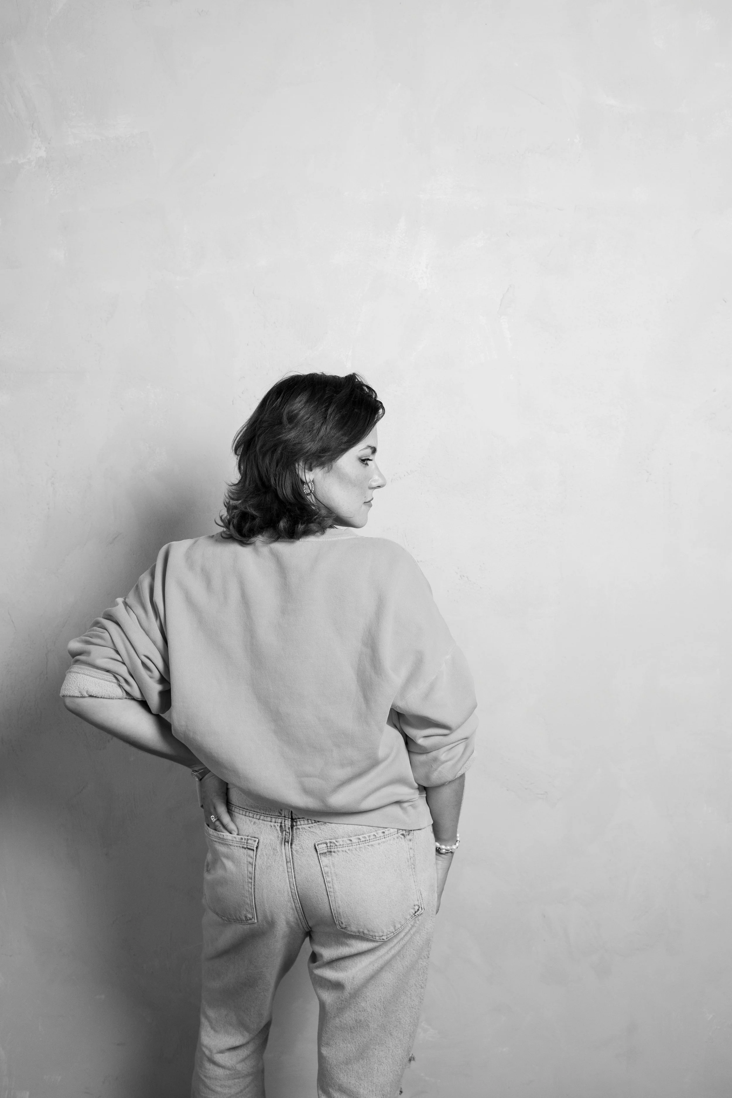 A woman with shoulder-length hair in a casual outfit, standing against a plain wall, seen from the back in black and white.