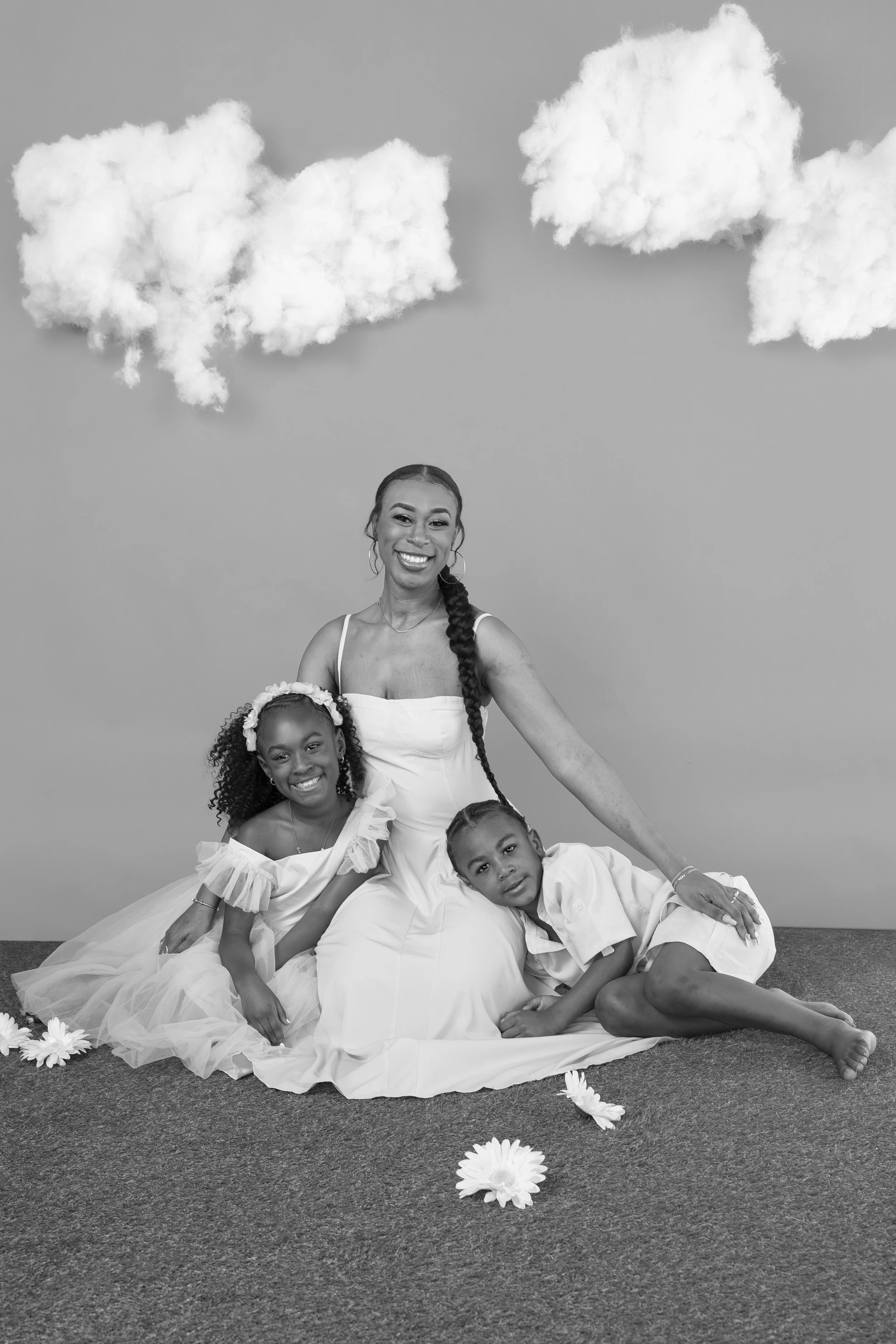 A woman and two children sitting on a carpeted floor with clouds made of cotton and flowers as decor.