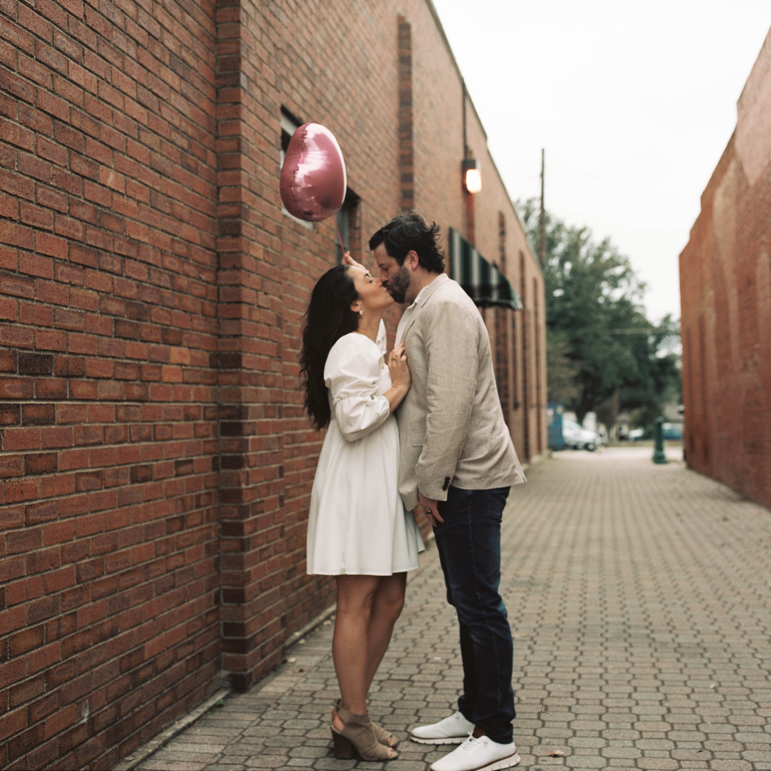A couple sharing a kiss on a city sidewalk, with a woman holding a pink heart-shaped balloon.