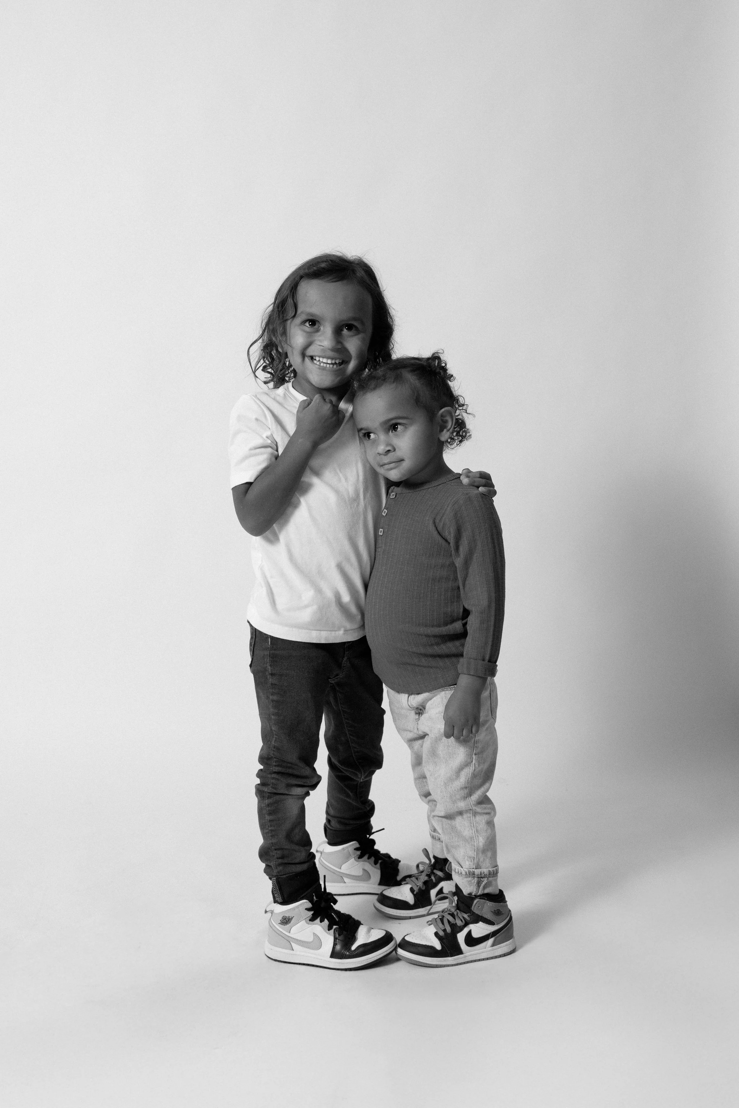 Two young girls standing together, one smiling with her arm around the other, both wearing sneakers and casual clothing, in a studio with a plain background.