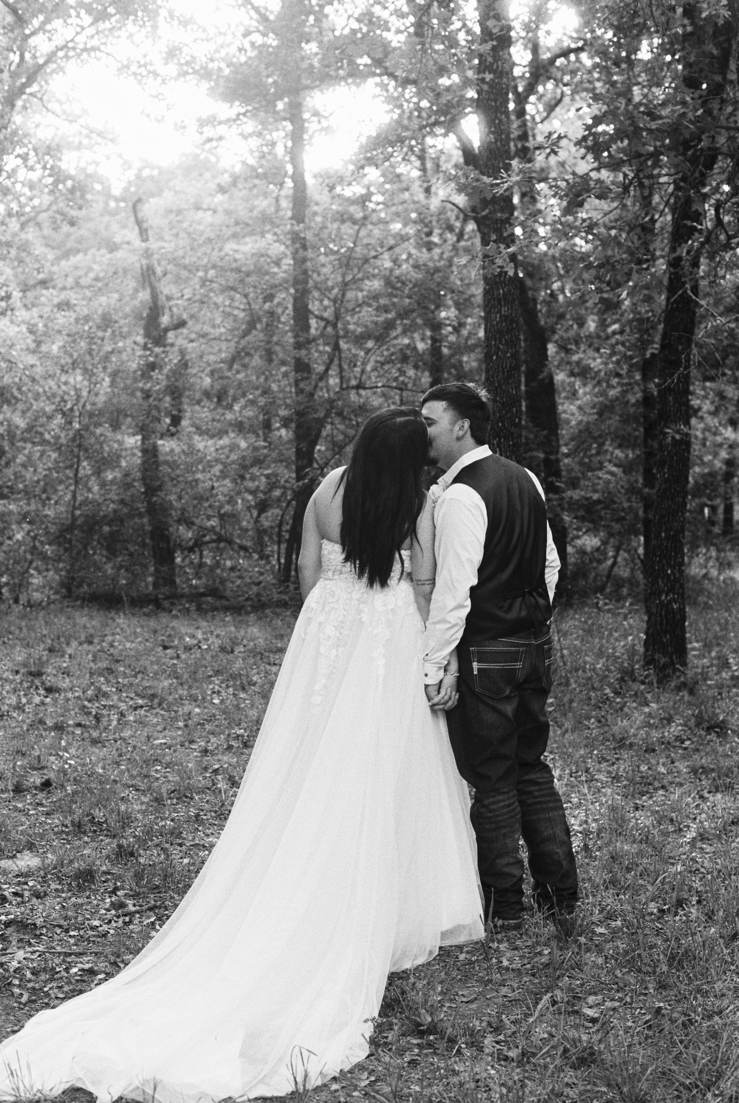 A couple dressed in wedding attire standing in a forest, kissing each other.