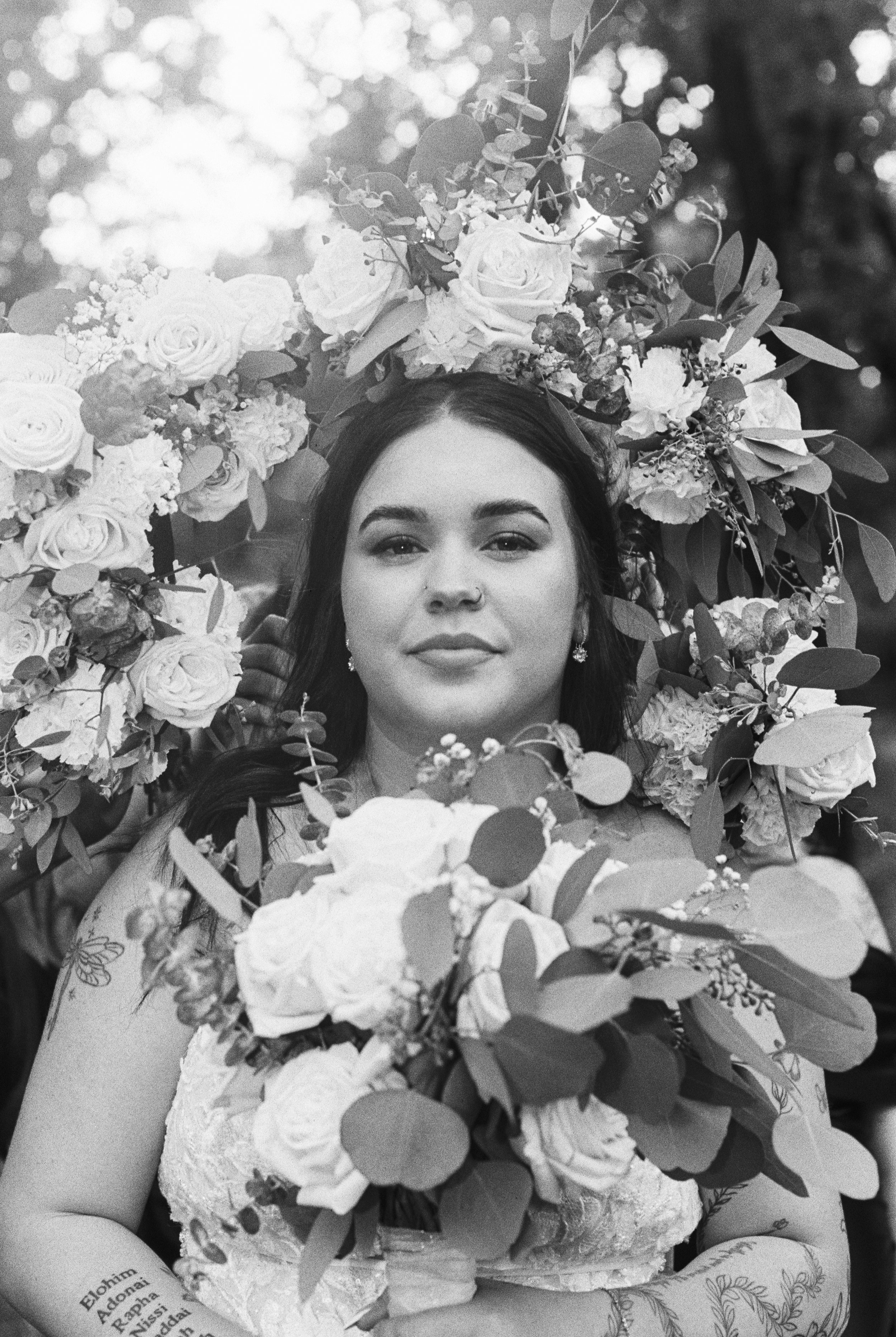 A woman with dark hair and tattoos on her arm, wearing a lace dress, holding a large bouquet of flowers, with a floral crown on her head, outdoors with sunlight and blurred background.