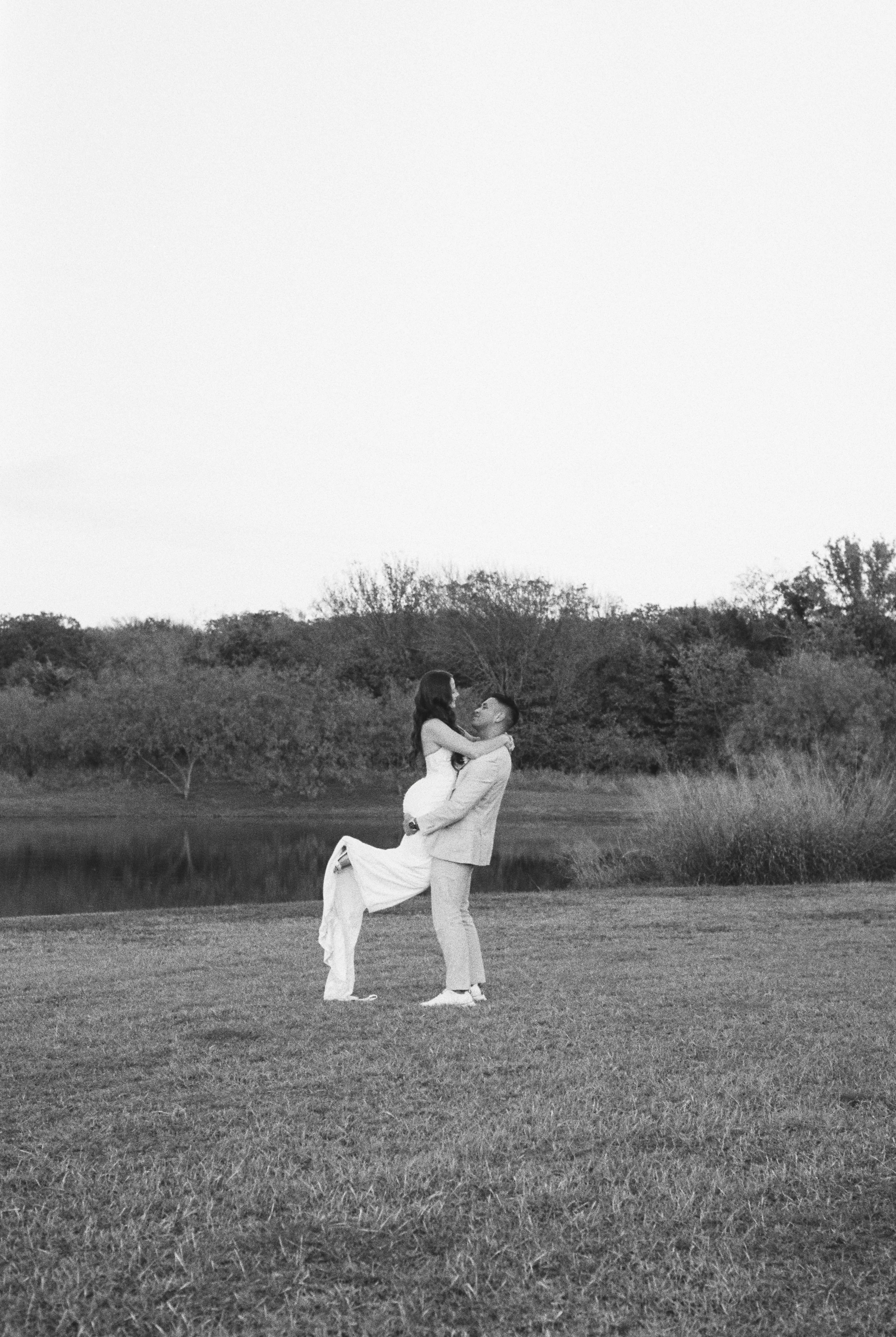 A couple, with the man lifting the woman, stands on a grassy field near a body of water with trees in the background, captured in black and white.
