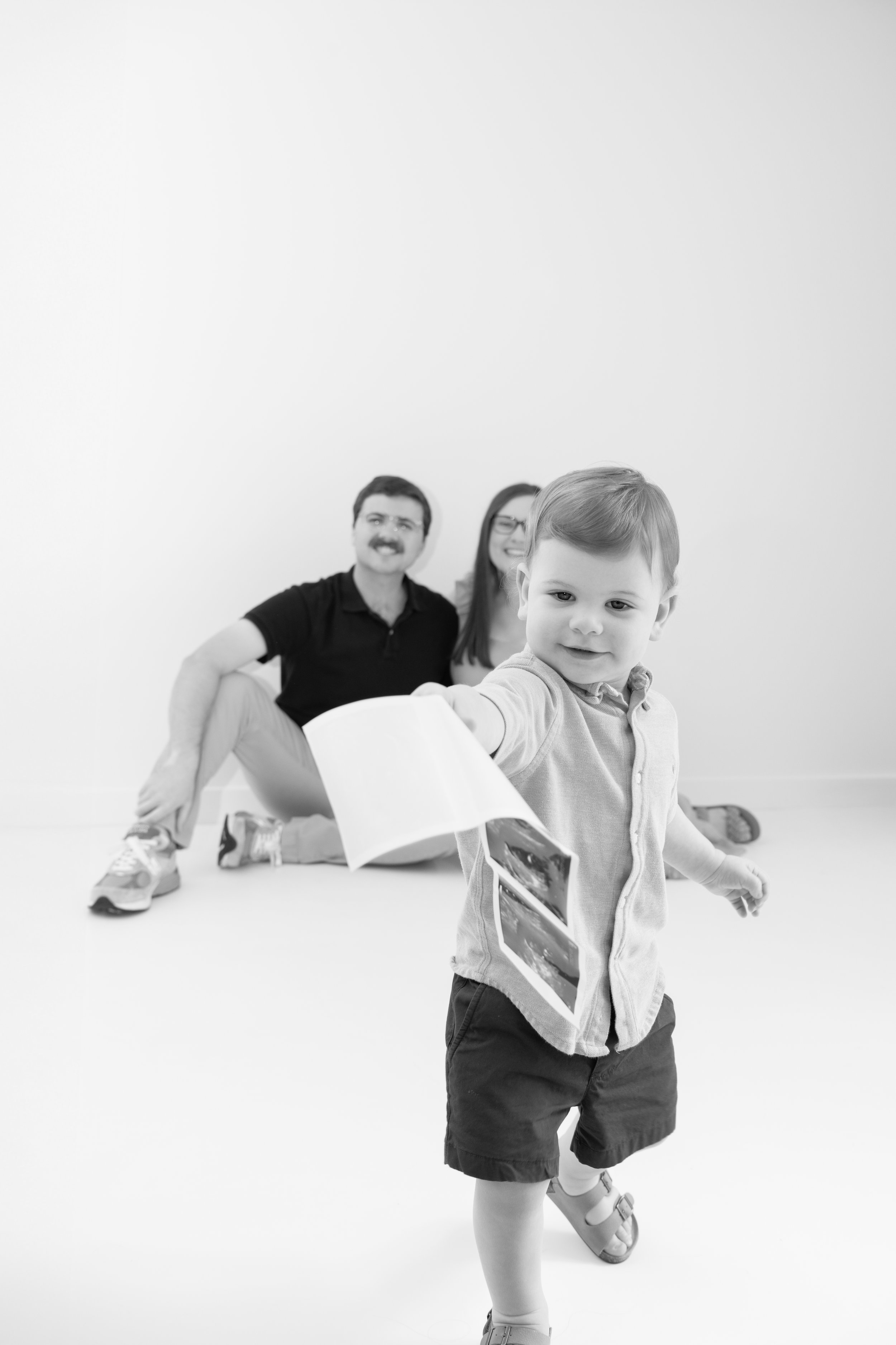 A young boy holding out photographs or postcards in the foreground, with a smiling family of three sitting on the floor in the background against a plain white wall.