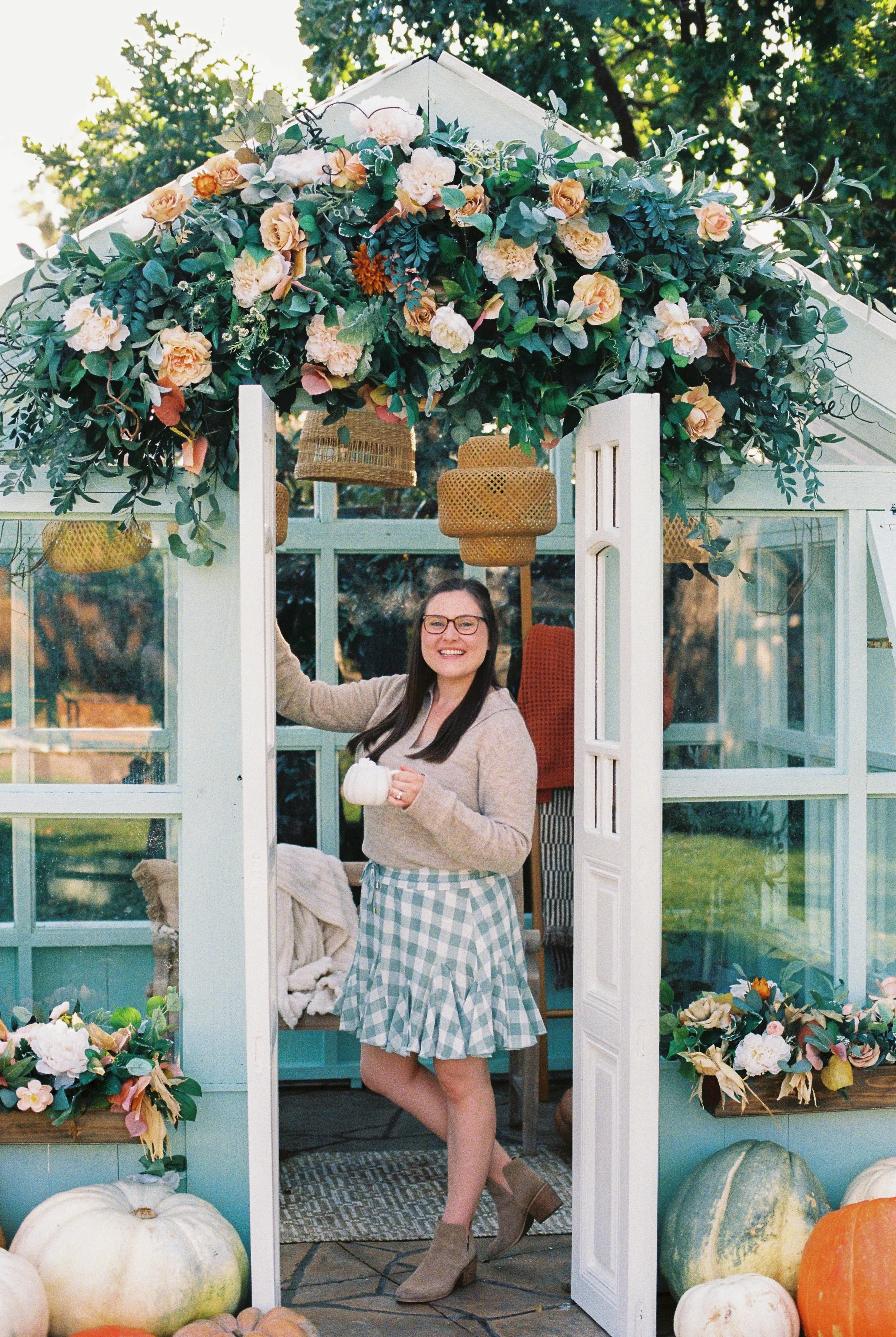 A woman standing in a decorated greenhouse with pumpkins and flowers, holding a small white pumpkin, smiling at the camera.