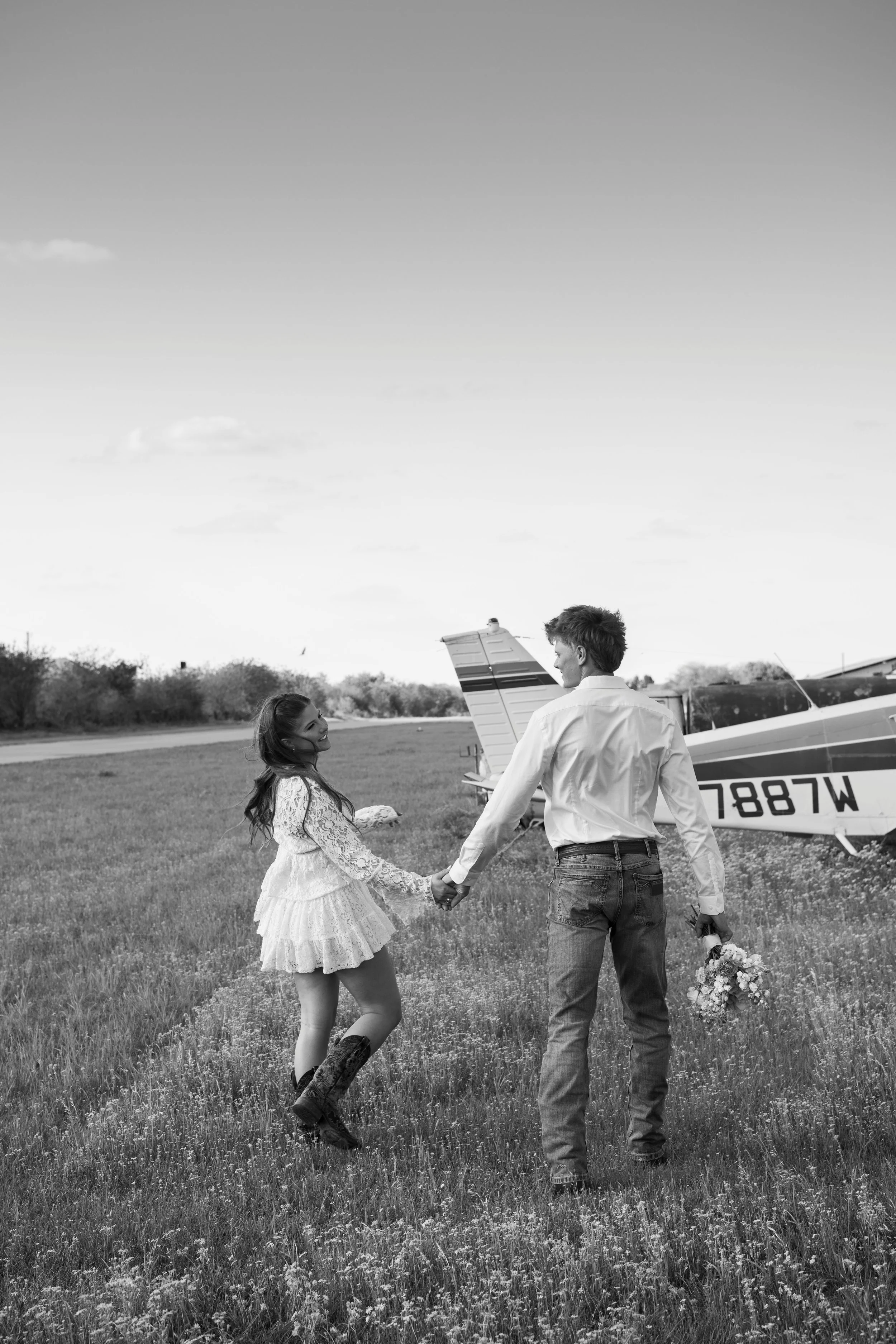 A black and white photo of a couple holding hands in a grassy field, with a small aircraft behind them and a clear sky overhead.