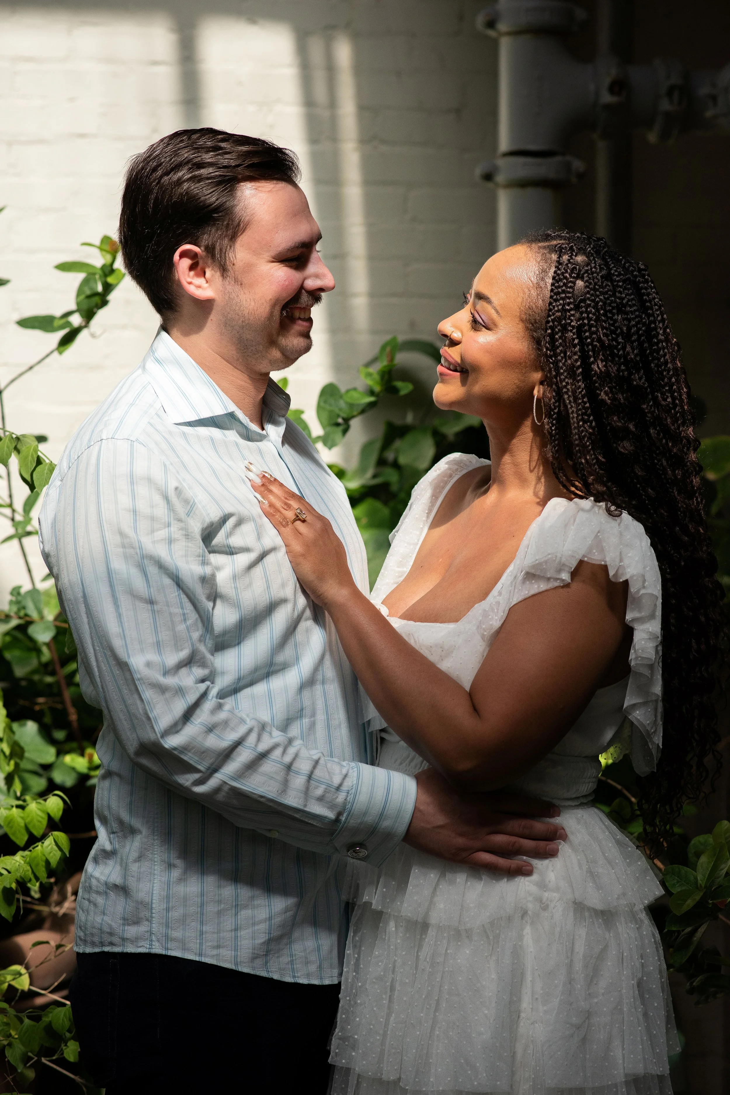 A happy couple stands close together, smiling at each other, outdoors near a brick wall and green plants.