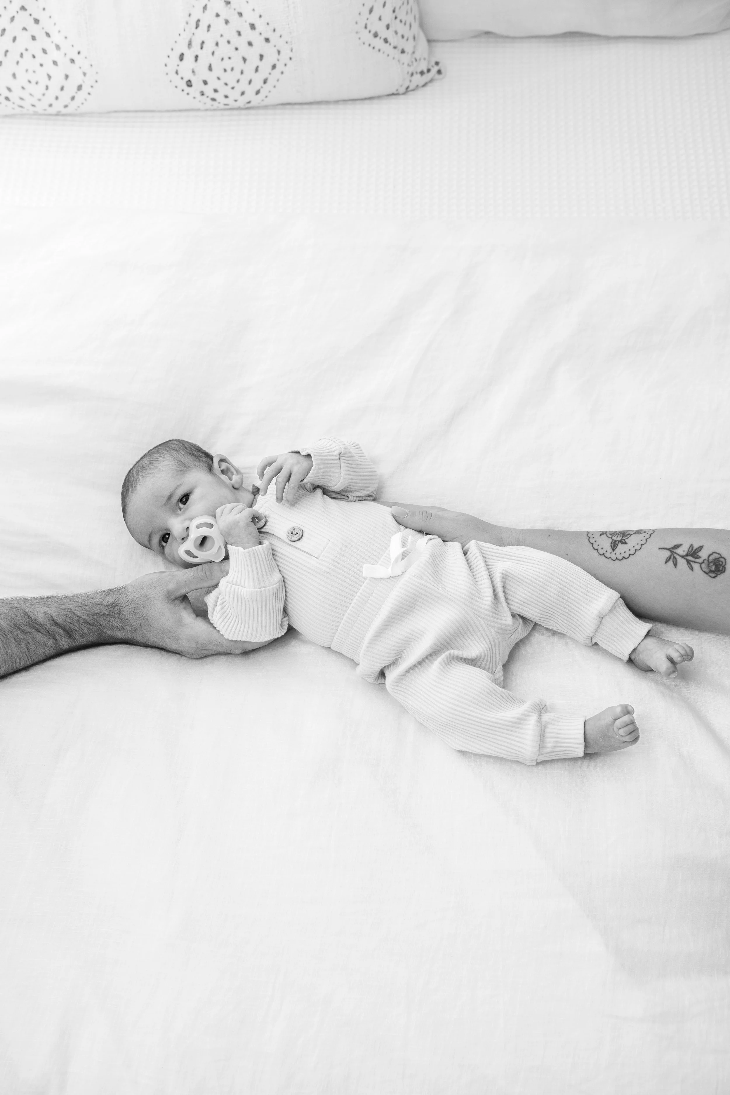 Black and white photo of a baby lying on a bed, with an adult's hand and arm near his head, and another arm supporting his body. The baby has a pacifier and is wearing a button-up shirt and pants.