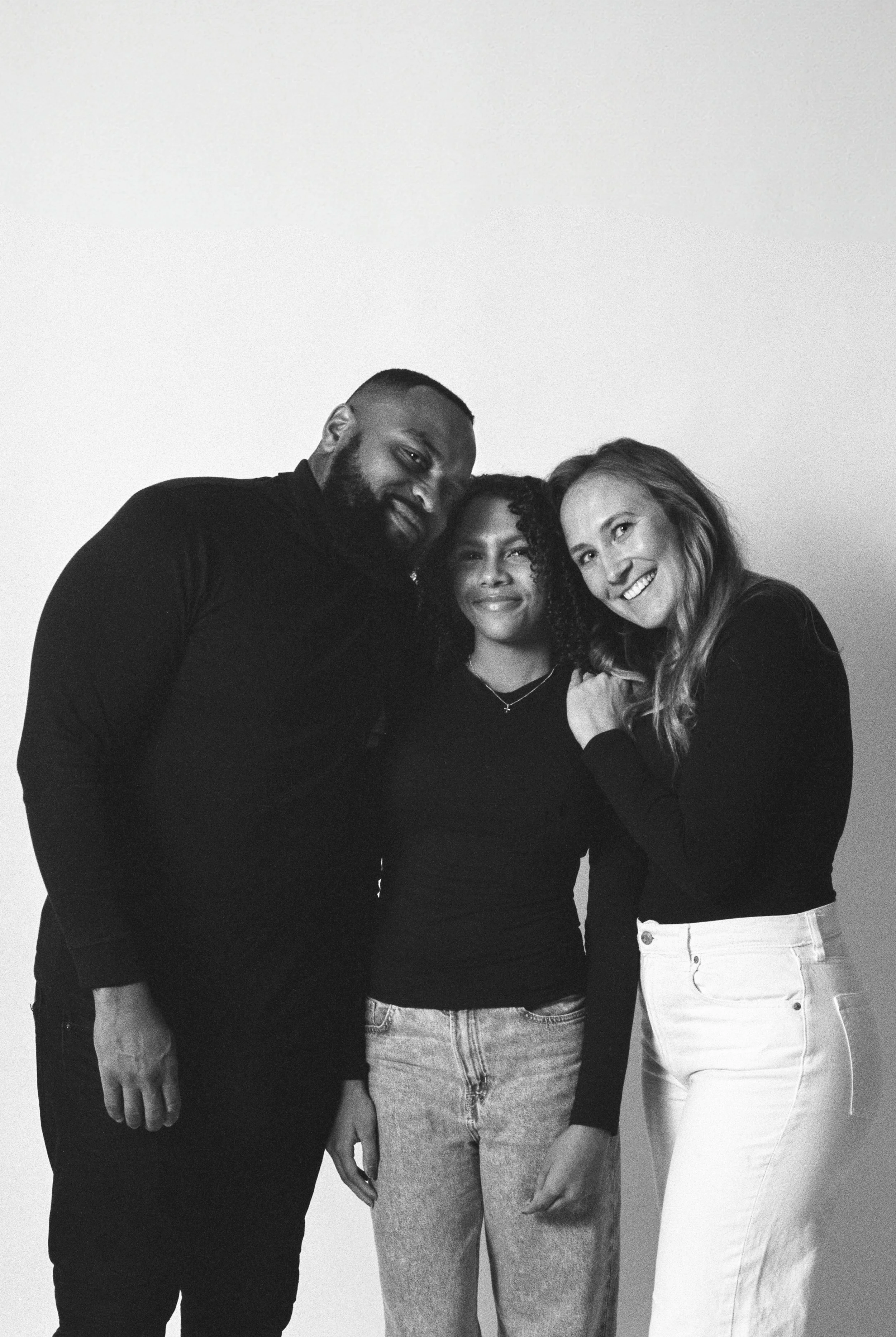 A black and white photo of a family of three standing close together in front of a plain wall, smiling at the camera.