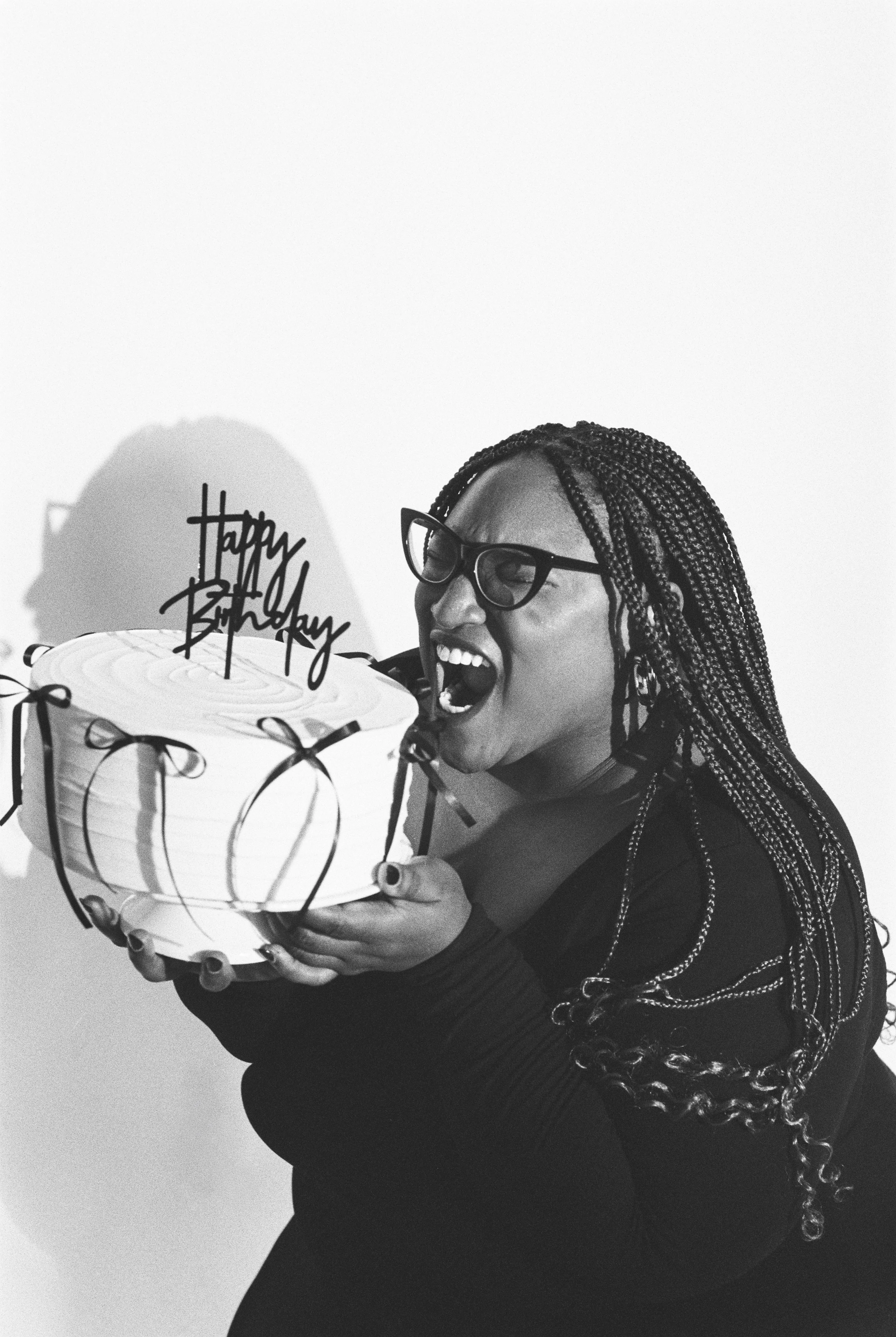 A woman with braided hair and glasses holding a birthday cake with a "Happy Birthday" topper, appearing to blow out candles.