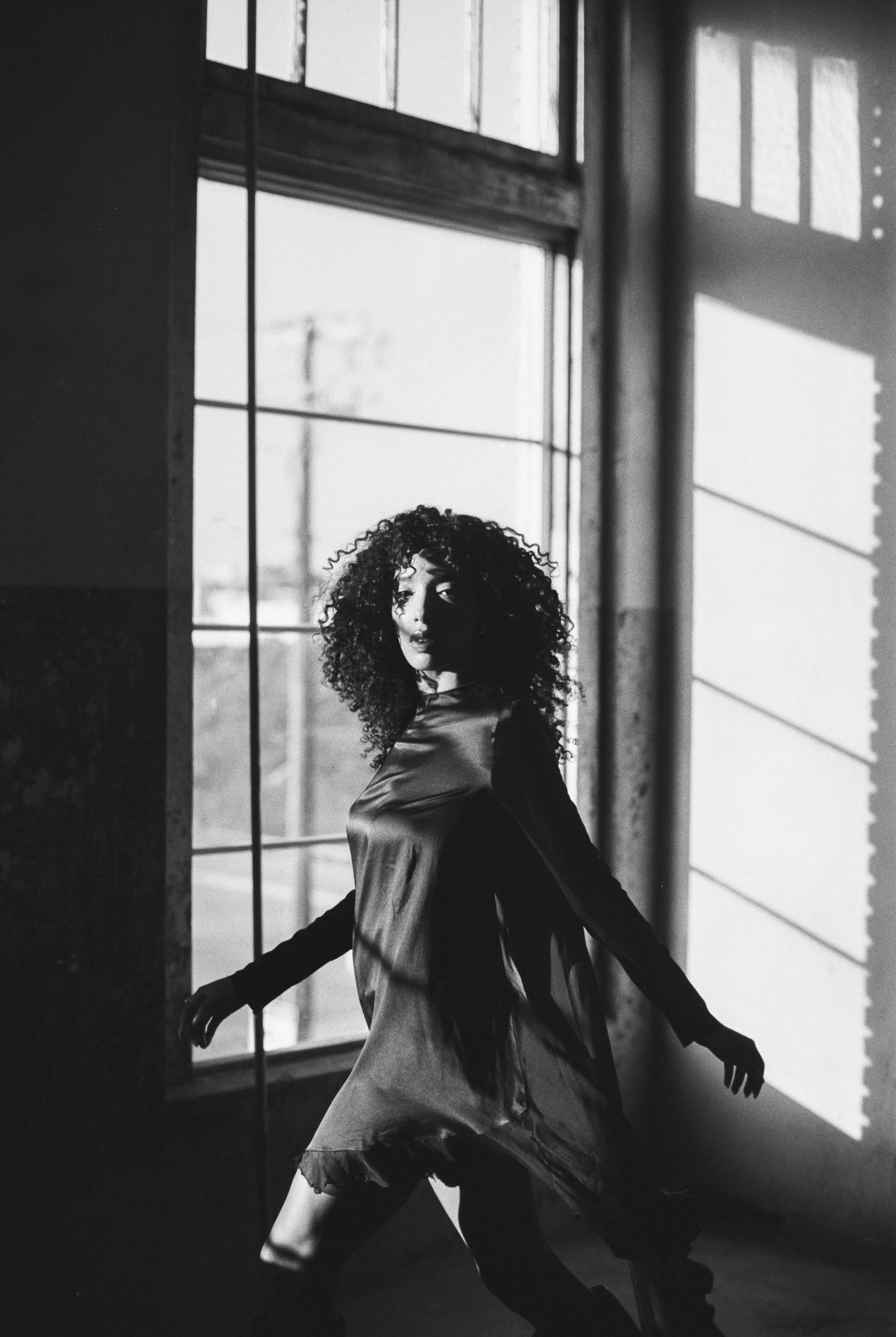A woman with curly hair in a long dress standing near a large window, with her arms extended outward. The scene is in black and white with sunlight casting shadows inside the room.
