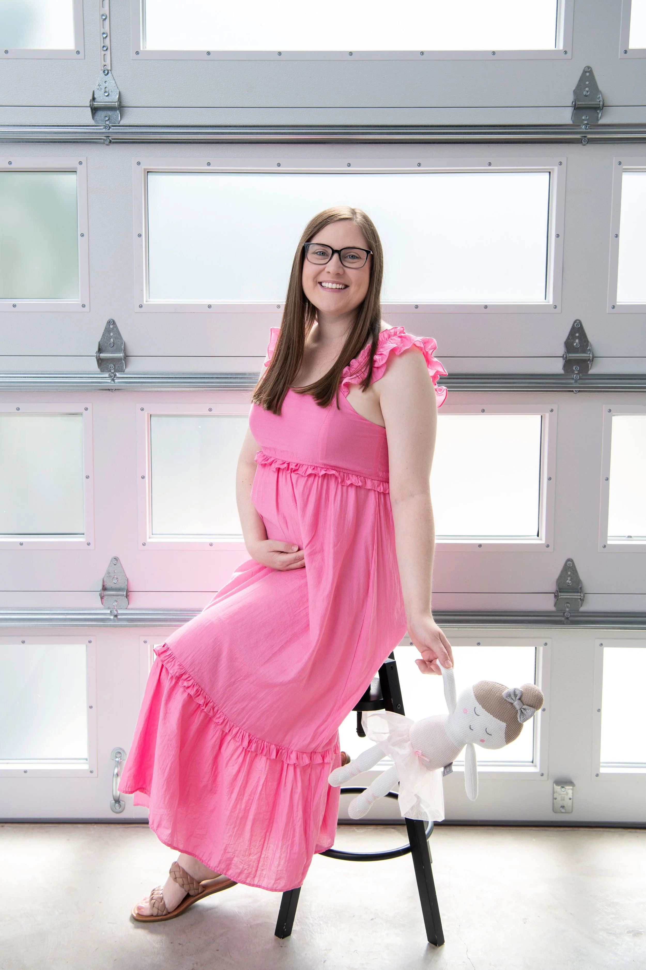 A woman wearing glasses and a long pink dress sits on a black stool holding a doll with a ballet tutu in front of a white garage door with windows.