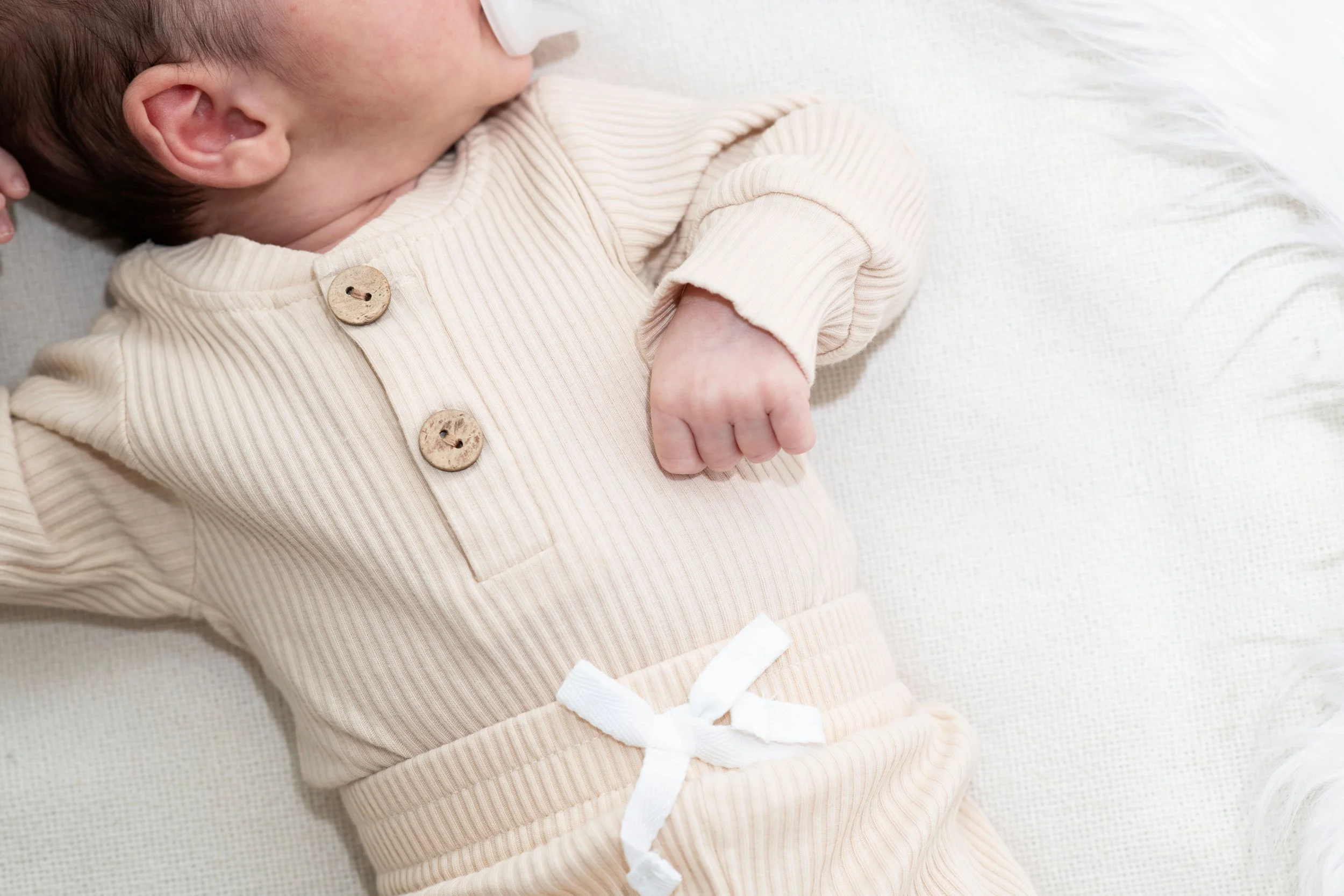 Close-up of a baby lying on a white surface, wearing a beige ribbed outfit with two wooden buttons and a white ribbon belt.