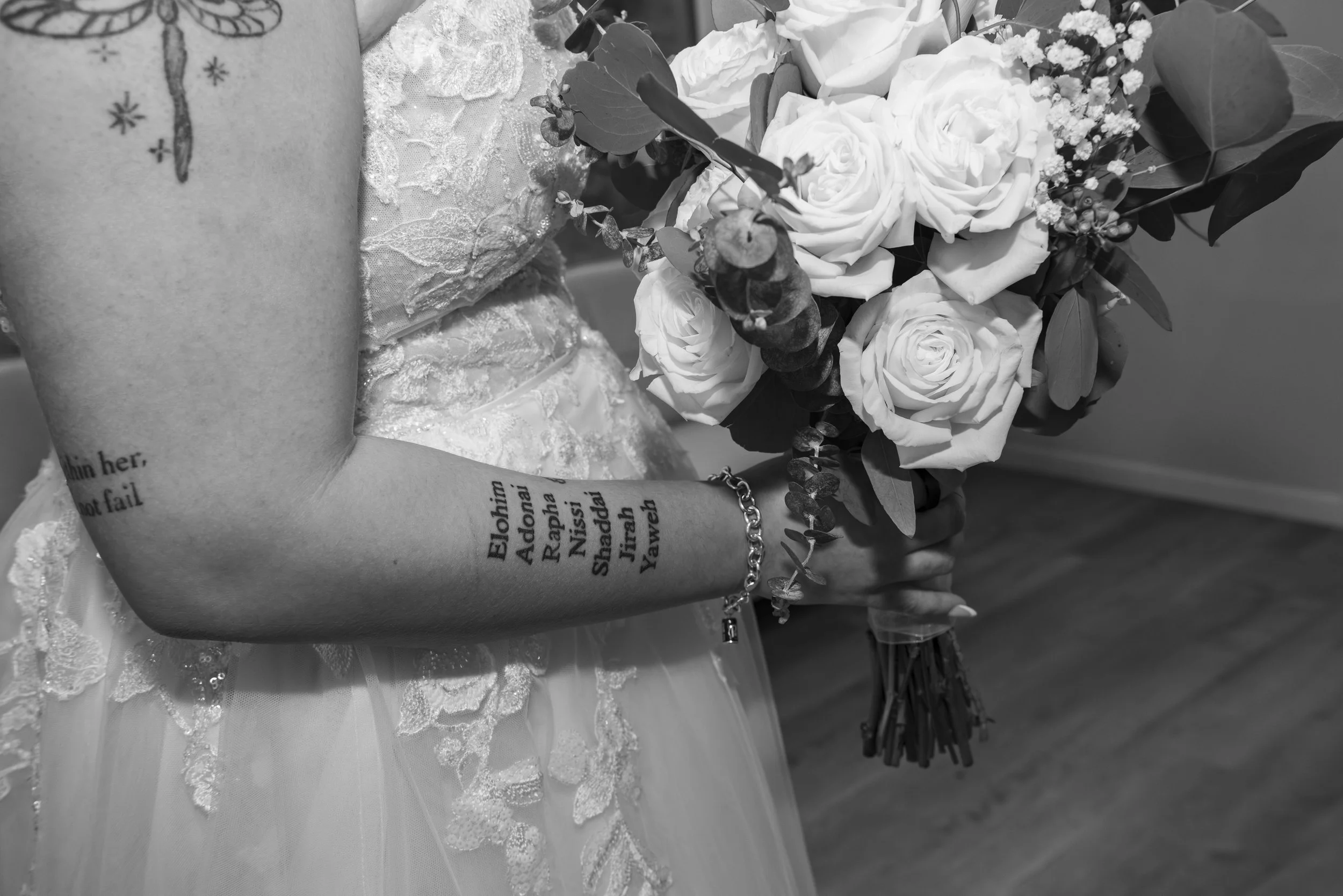 Close-up of a woman's arm with a tattoo and a bracelet, holding a bouquet of white roses and small flowers, with text on the tattoo visible.