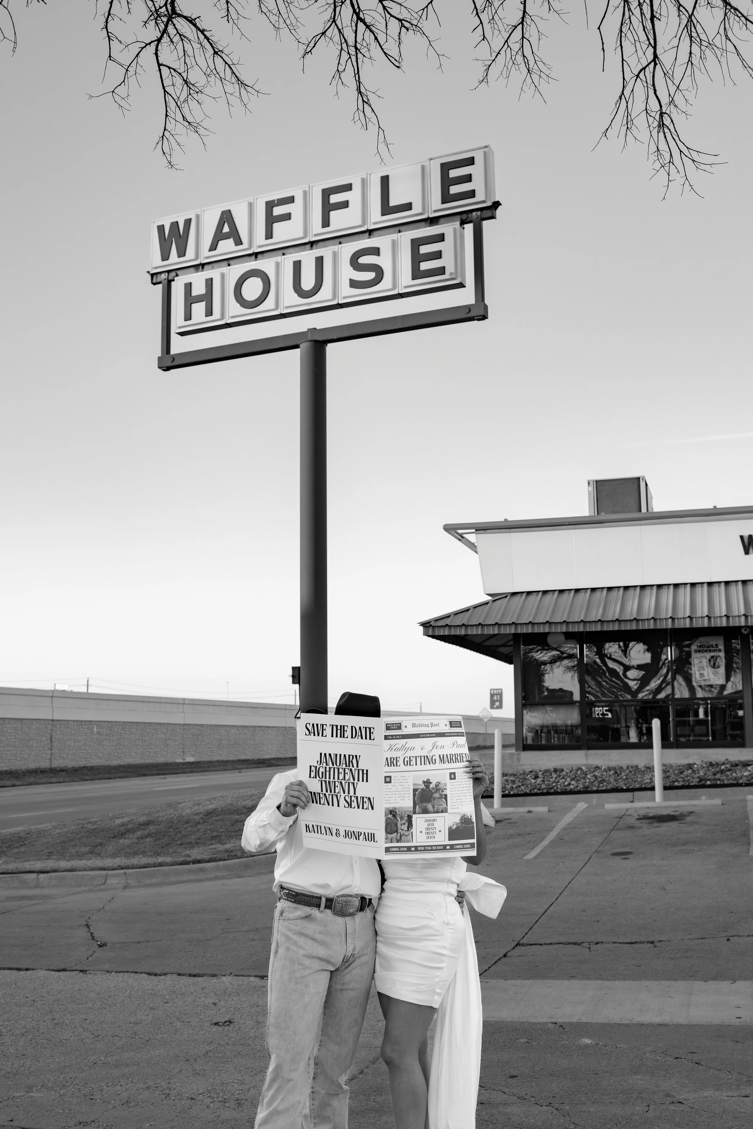 Two people are standing outside a restaurant holding up a newspaper with an announcement about a wedding. The restaurant sign reads 'WAFFLE HOUSE' and is mounted on a pole against a cloudy sky. The scene appears to be in a parking lot with a building