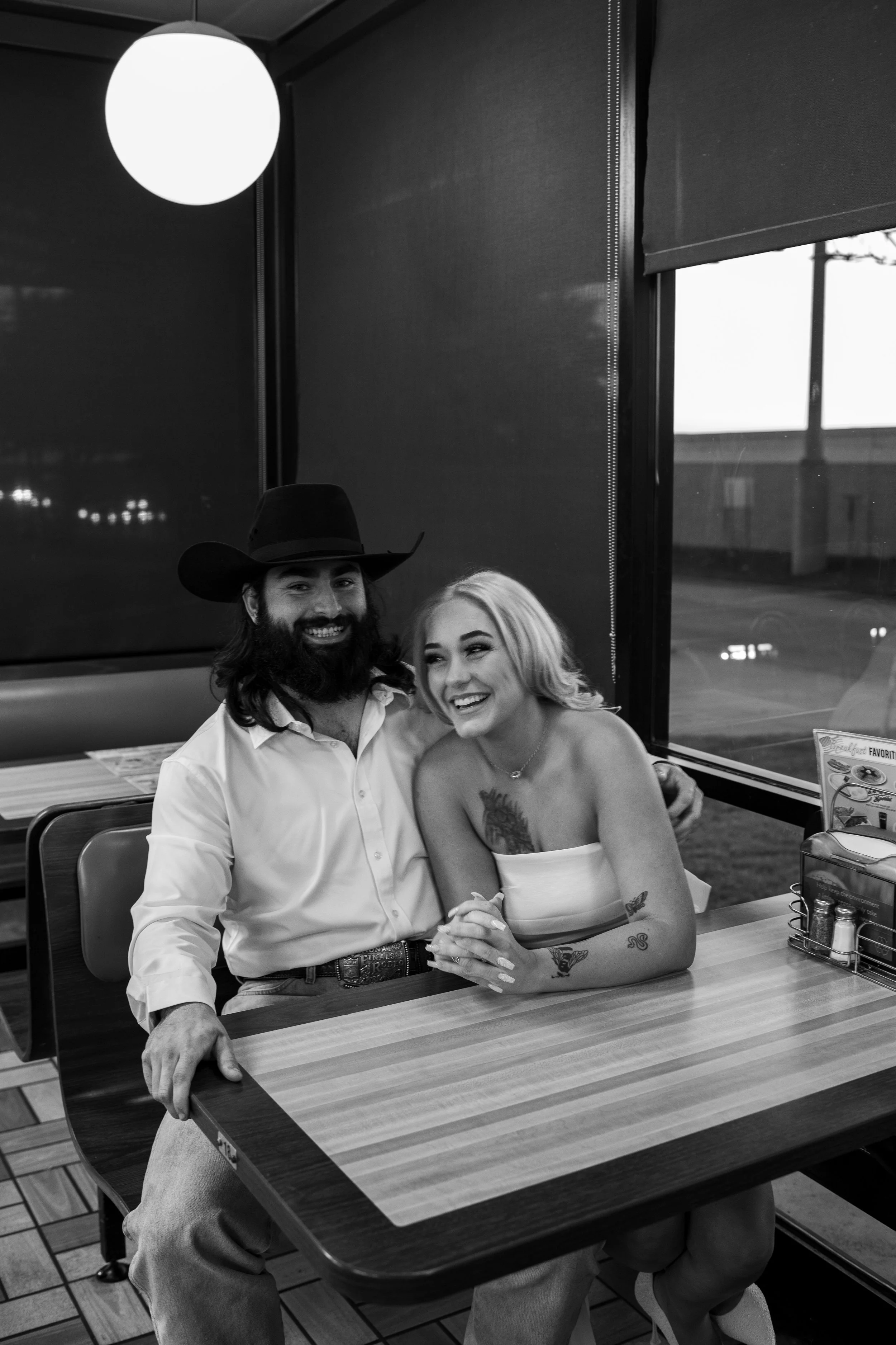 A happy man with a beard, wearing a cowboy hat and a white shirt, sitting next to a smiling woman with blonde hair, tattoos on her arms, and wearing a strapless top, inside a diner. They are holding hands on a table with condiments and a menu, and ha