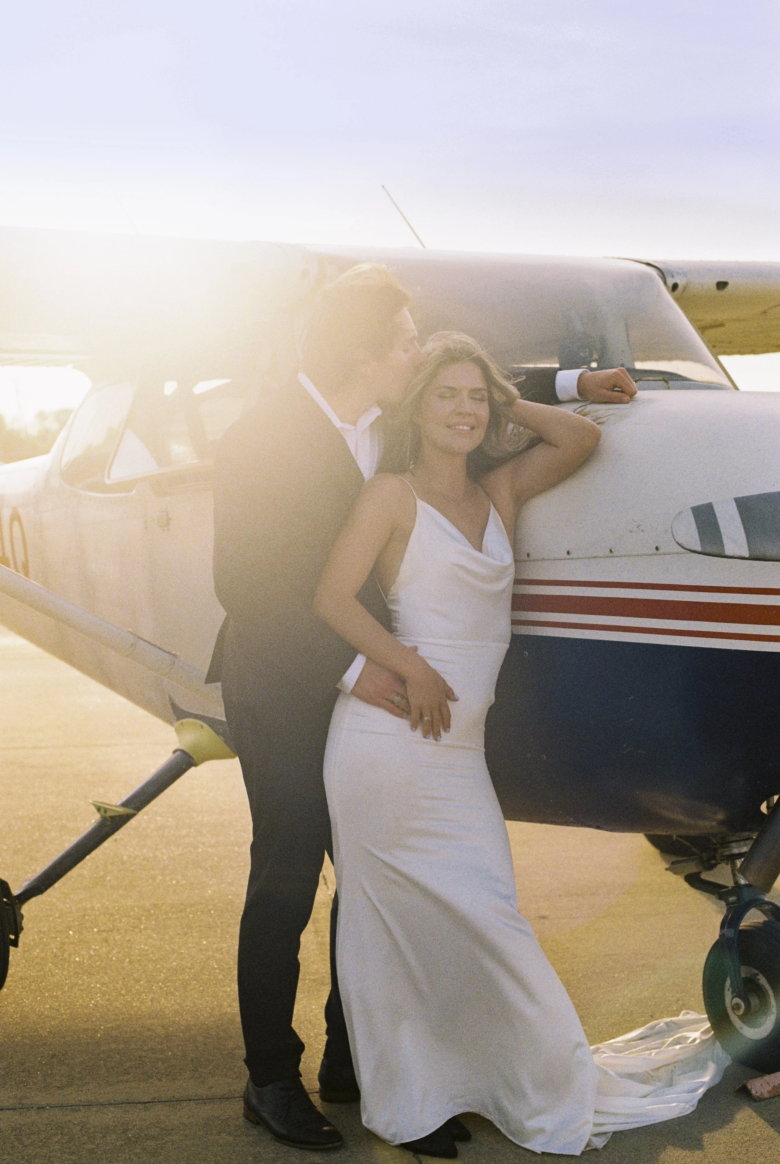 A bride in a white wedding dress stands beside a pilot in a suit, smiling as they pose in front of a small airplane during what appears to be golden hour, with sunlight creating a warm glow.